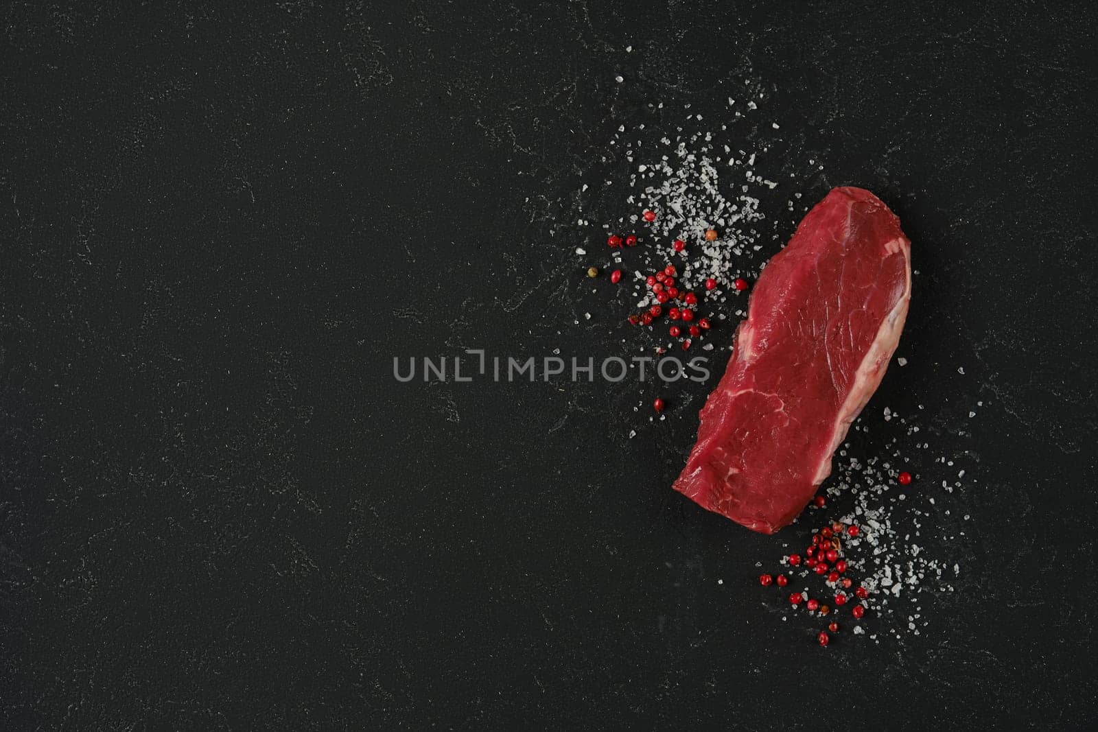 A piece of fresh raw beef sirloin steak on a dark countertop surrounded by coarse sea salt and red peppercorns, showcasing a clean and elegant presentation ideal for meal preparation.