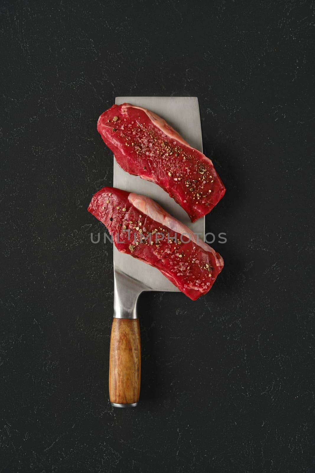 Two raw beef steaks seasoned with spices on a large metal cleaver with a wooden handle. The dark countertop highlights the vibrant red color of the meat, indicating freshness.