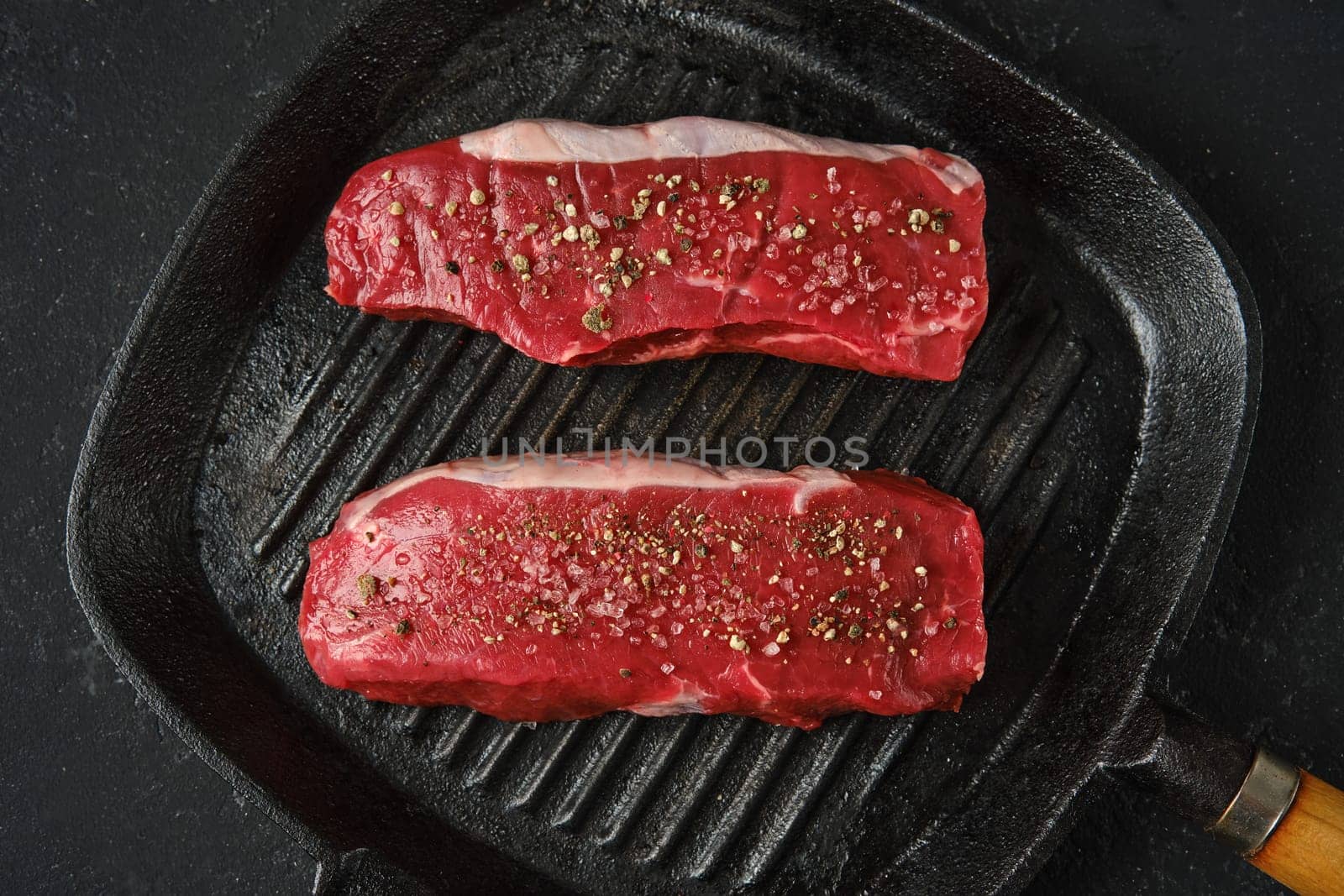 Top view of two thick cuts of raw beef steaks on a cast iron skillet. The marbled texture of the meat is seasoned with sea salt and pepper