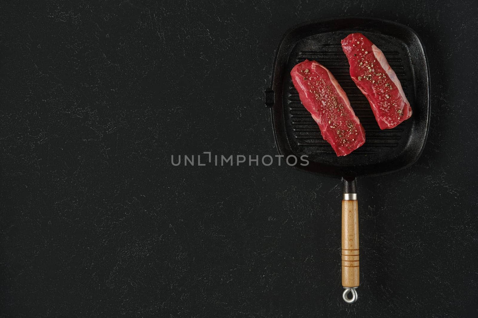 Two raw beef steaks on a preheated griddle pan, seasoned with spices, in a home kitchen setting. The black surface is for text