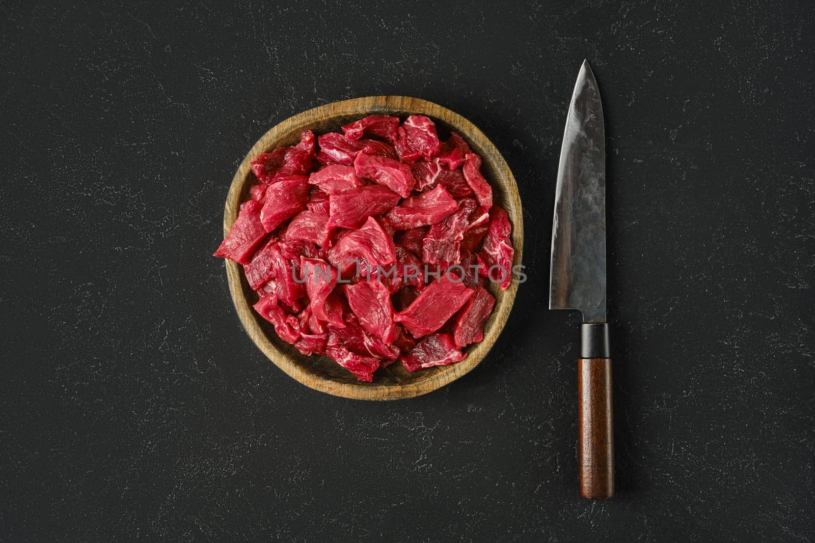 A wooden bowl holds freshly cut pieces of raw beef, showcasing a vibrant red color. Beside the bowl, a sharp knife rests on a dark textured surface, ready for preparation or cooking.