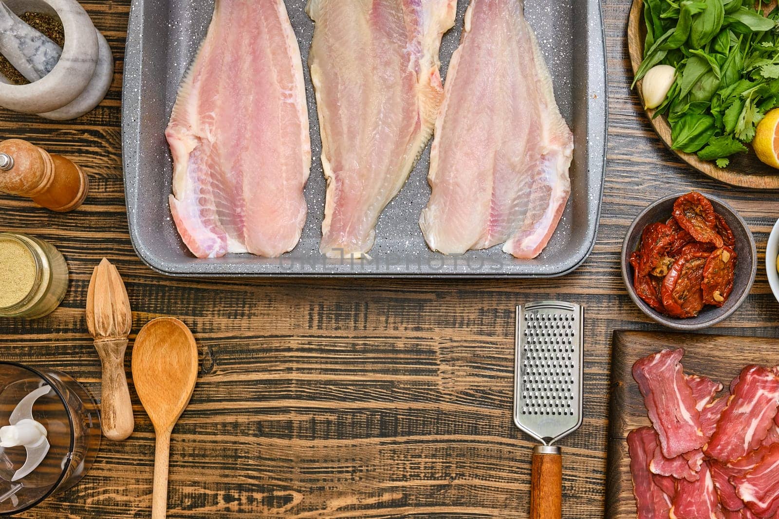 Fresh fish fillets are laid out on a baking tray, surrounded by various ingredients including herbs, lemon, and spices. Wooden utensils and bowls add a rustic touch to the kitchen workspace.