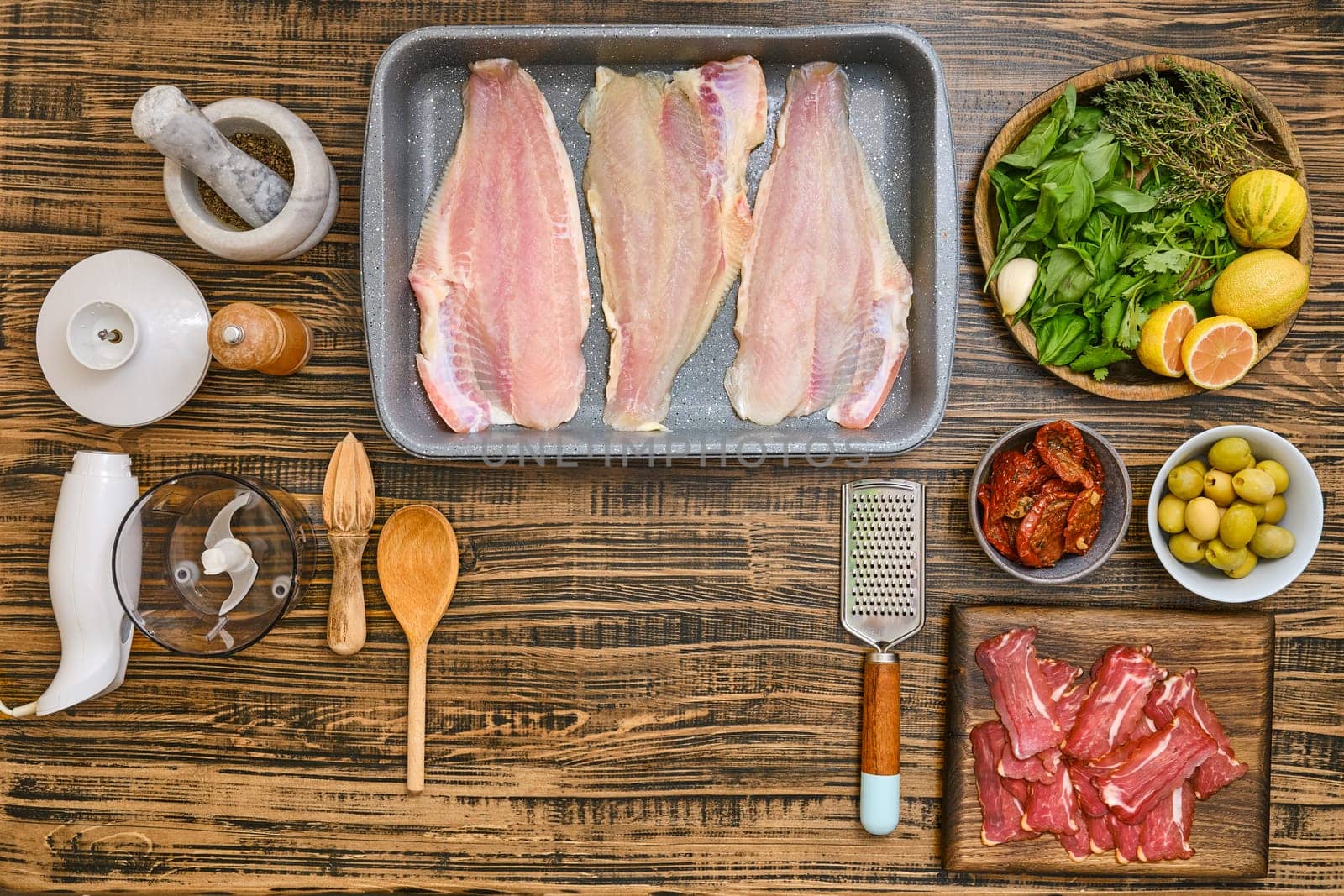 Various fresh ingredients are arranged on a wooden tabletop, including fish fillets, herbs, olives, and spices. This setup is perfect for creating a delightful seafood meal.