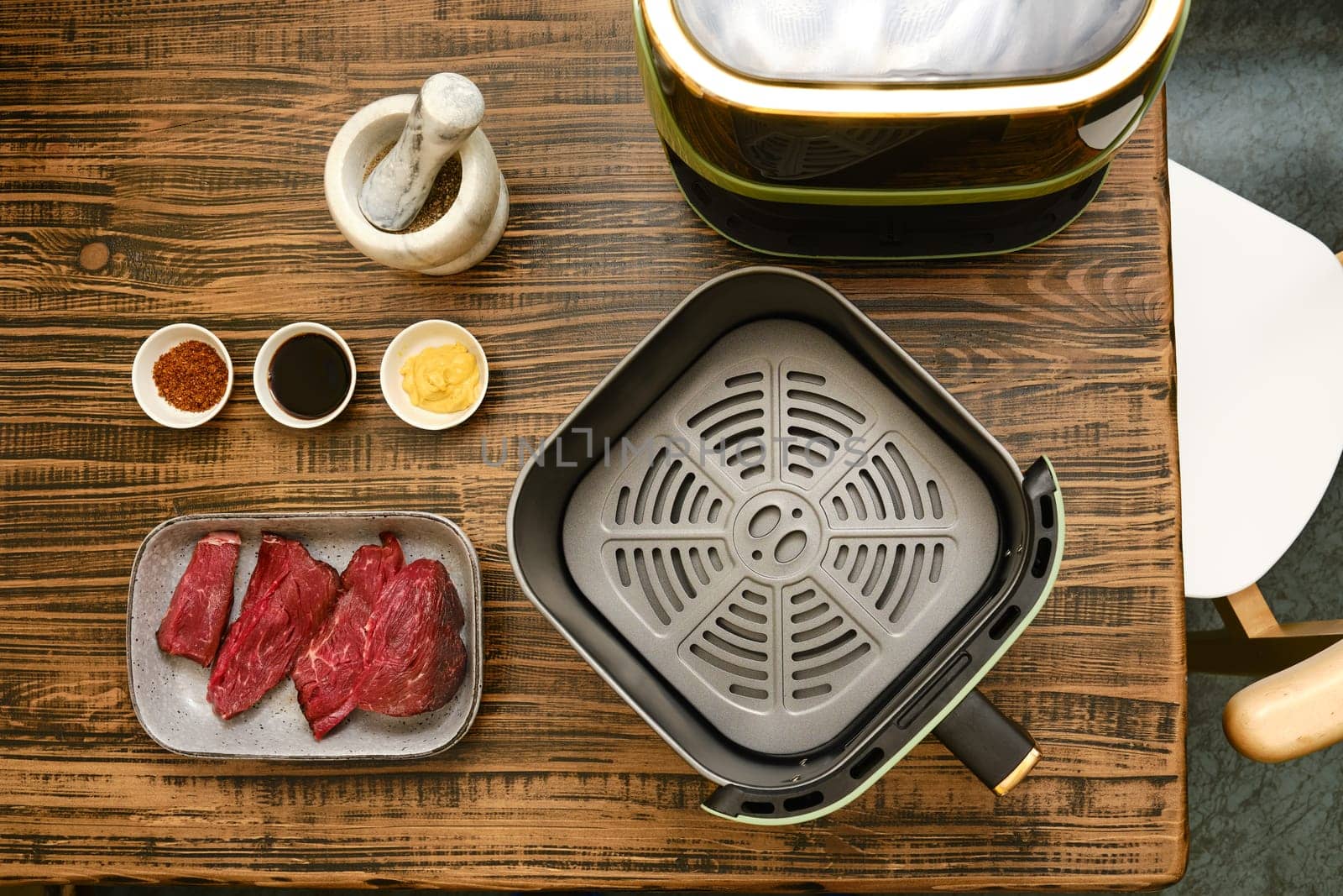 Fresh cuts of meat are arranged alongside several bowls of spices and sauces on a wooden table. A cooking appliance sits nearby, ready for a grilling or air-frying session.