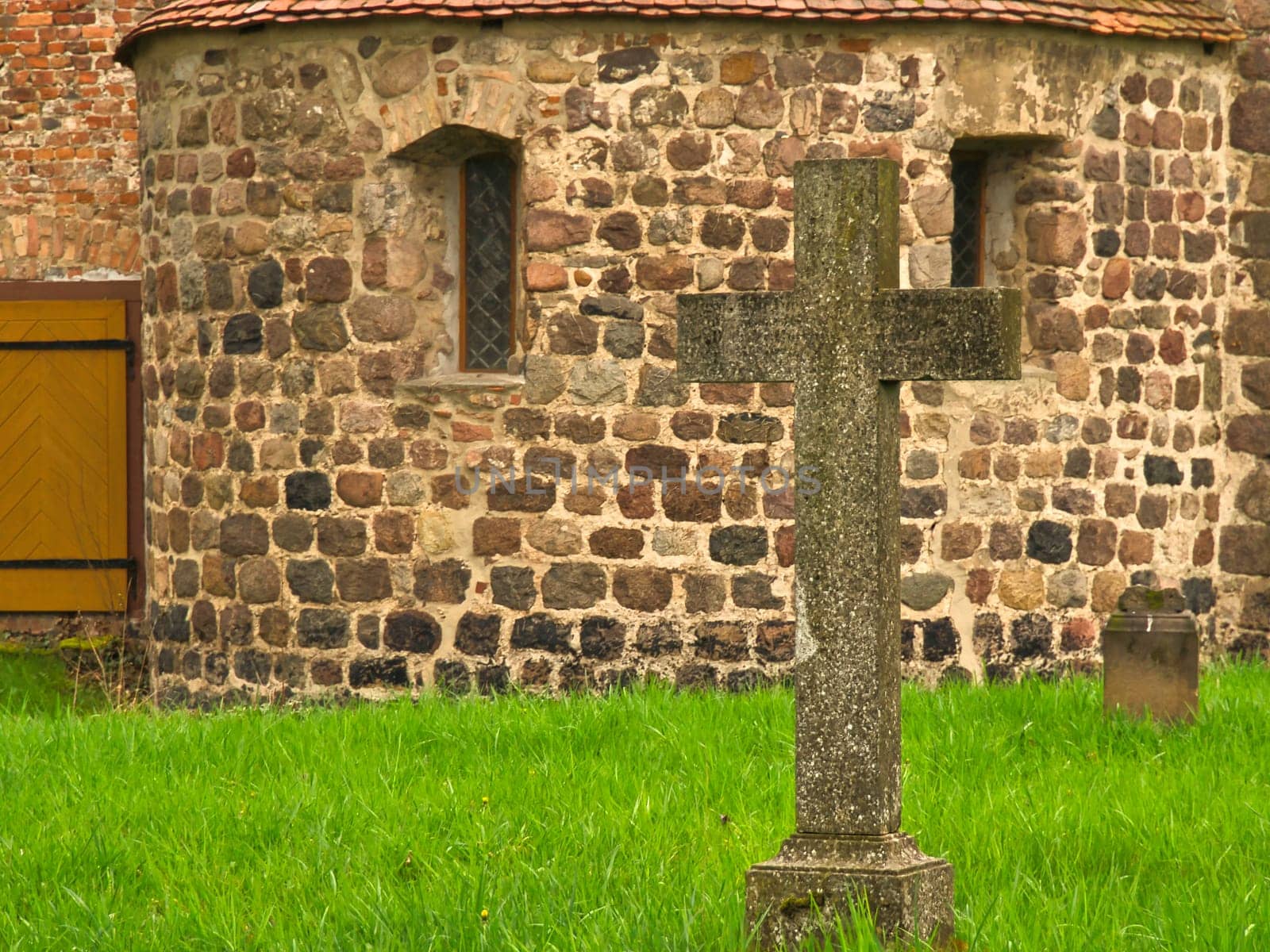 A historic stone cross in front of an old rustic church with small windows. Lively green grass in the foreground adds depth and contrast to the picture.