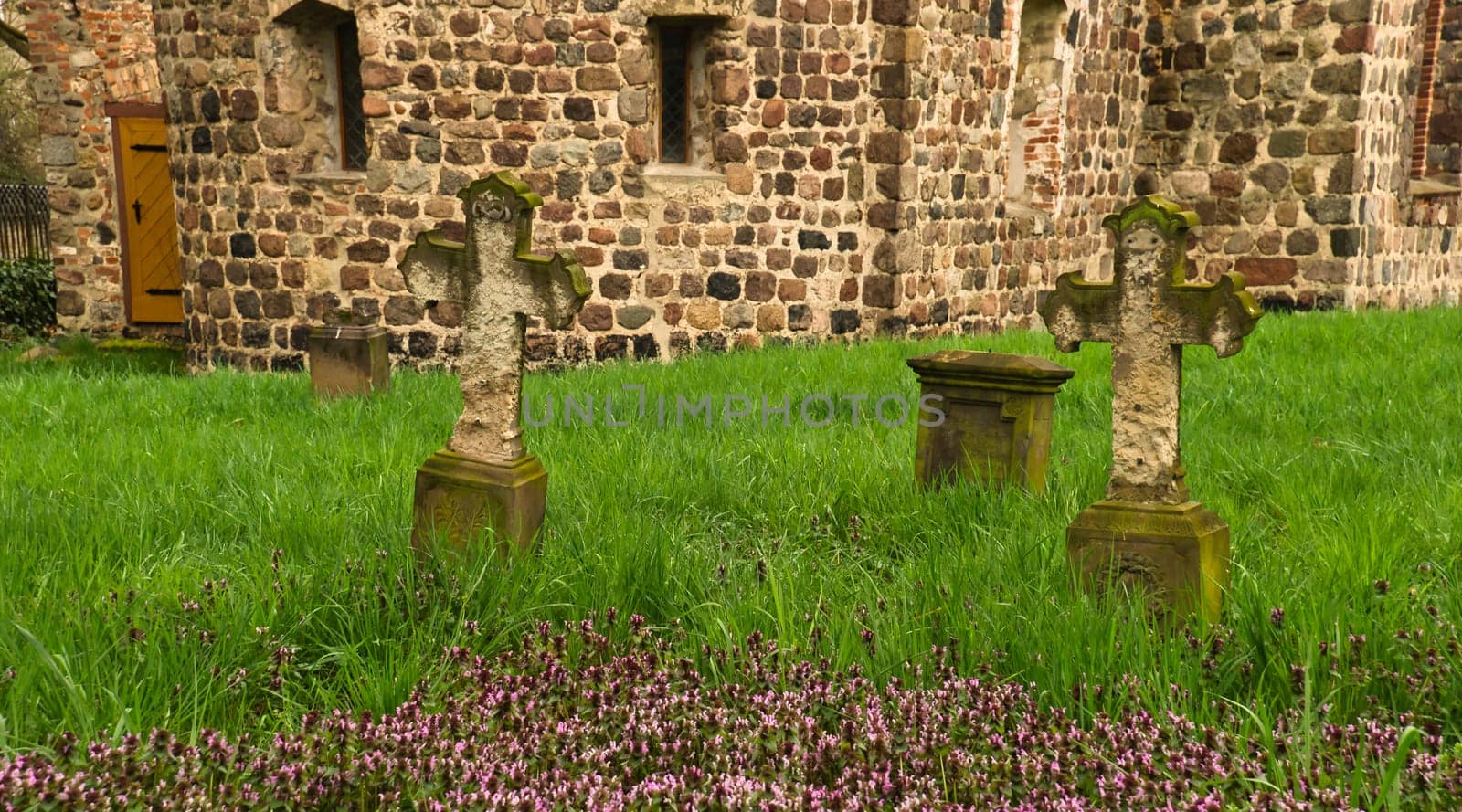 A historic stone cross in front of an old rustic church with small windows. Lively green grass in the foreground adds depth and contrast to the picture.