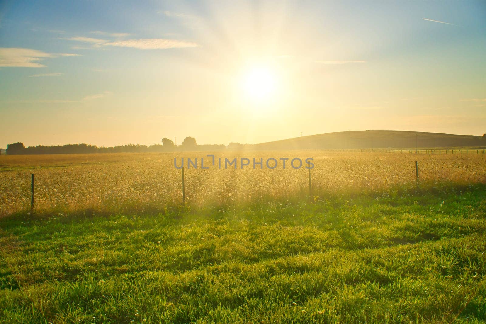 Sunset over a wide field with rolling hills in the background, a wooden fence in the foreground. Warm mood lighting and silhouettes emphasize the landscape.
