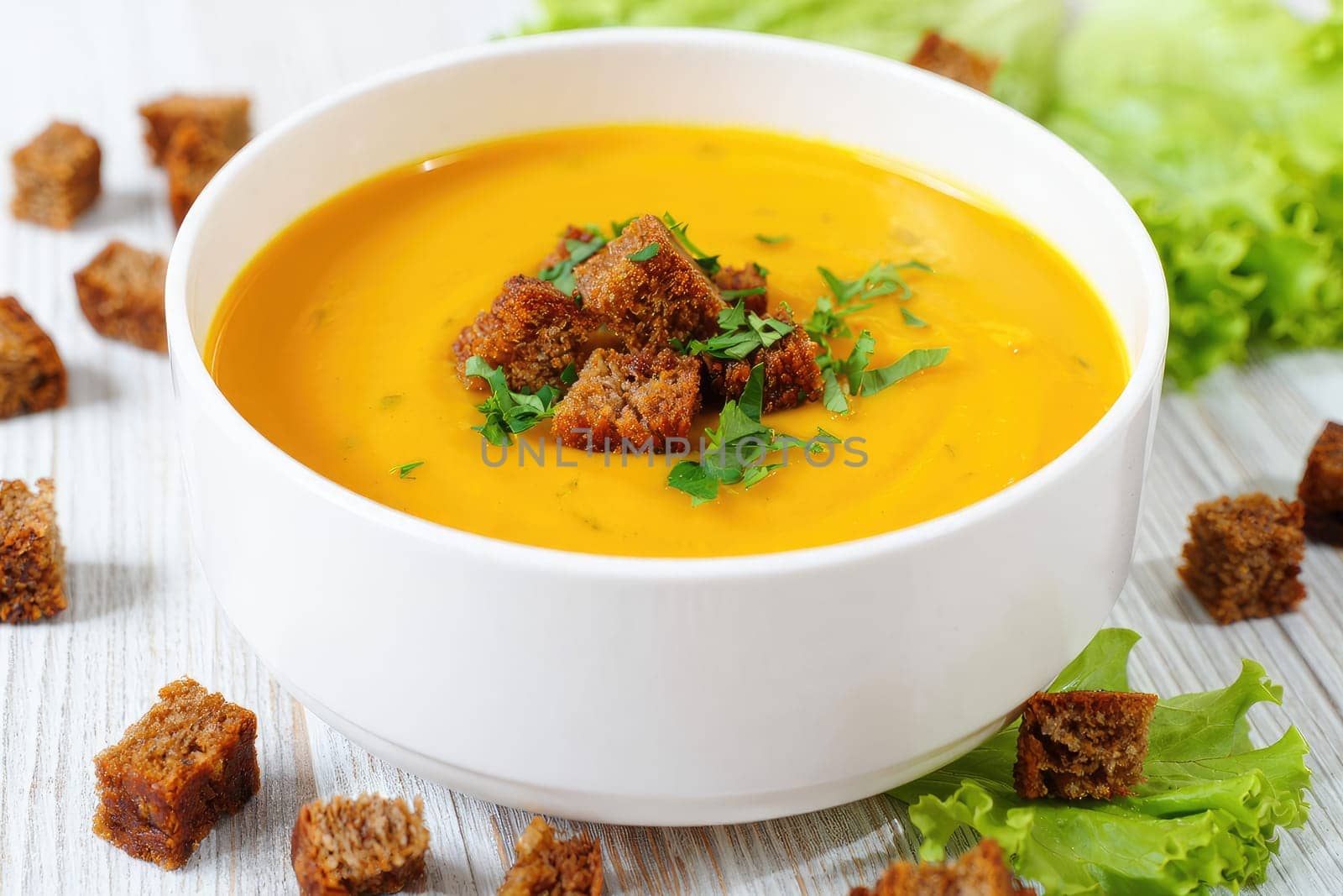 A warm bowl of creamy pumpkin soup topped with crunchy croutons sits on a wooden surface. Fresh greens surround the bowl, enhancing the appetizing appearance of the dish.