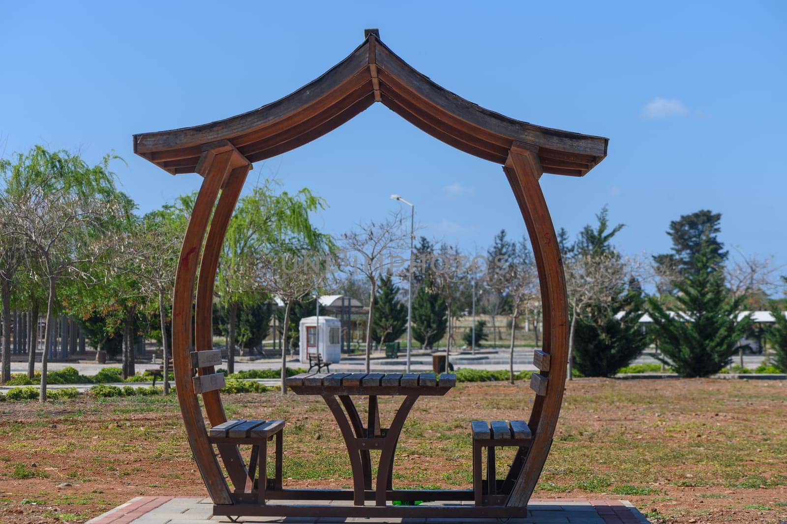 A wooden gazebo stands invitingly in a sunny park, surrounded by lush greenery and gentle trees. The clear blue sky enhances the serenity, making it a perfect place to unwind.