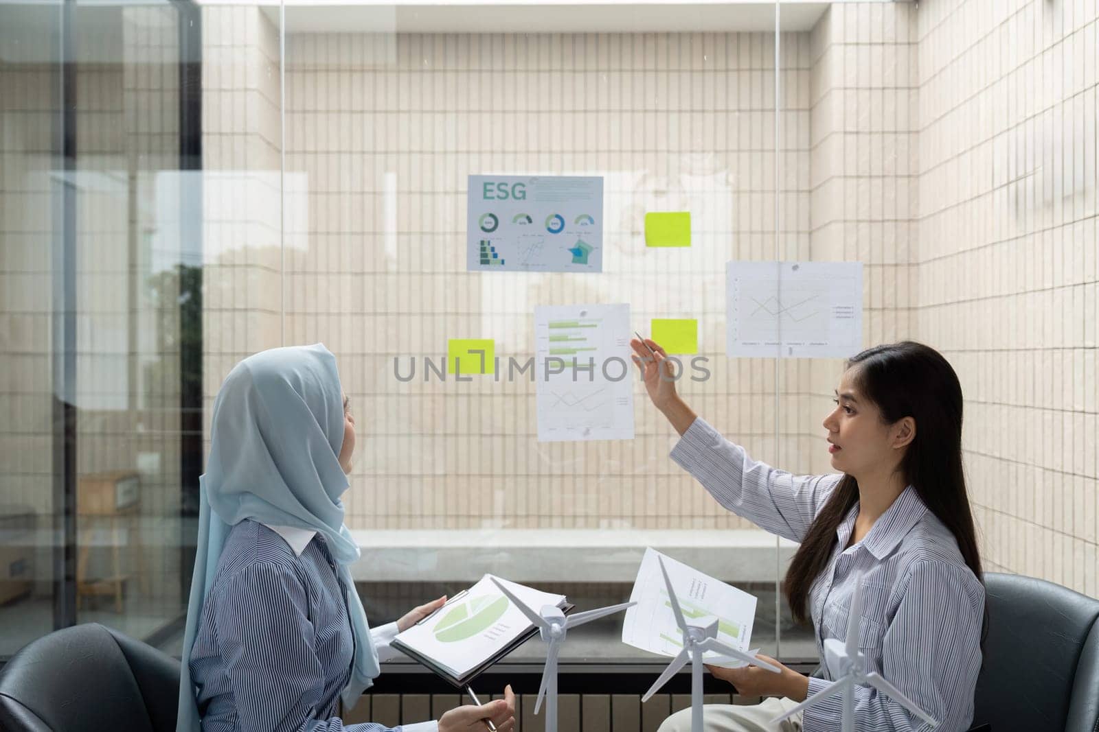 In a glass-walled office, two women engage in a dynamic discussion about ESG initiatives, analyzing data and sharing insights to enhance sustainable practices.