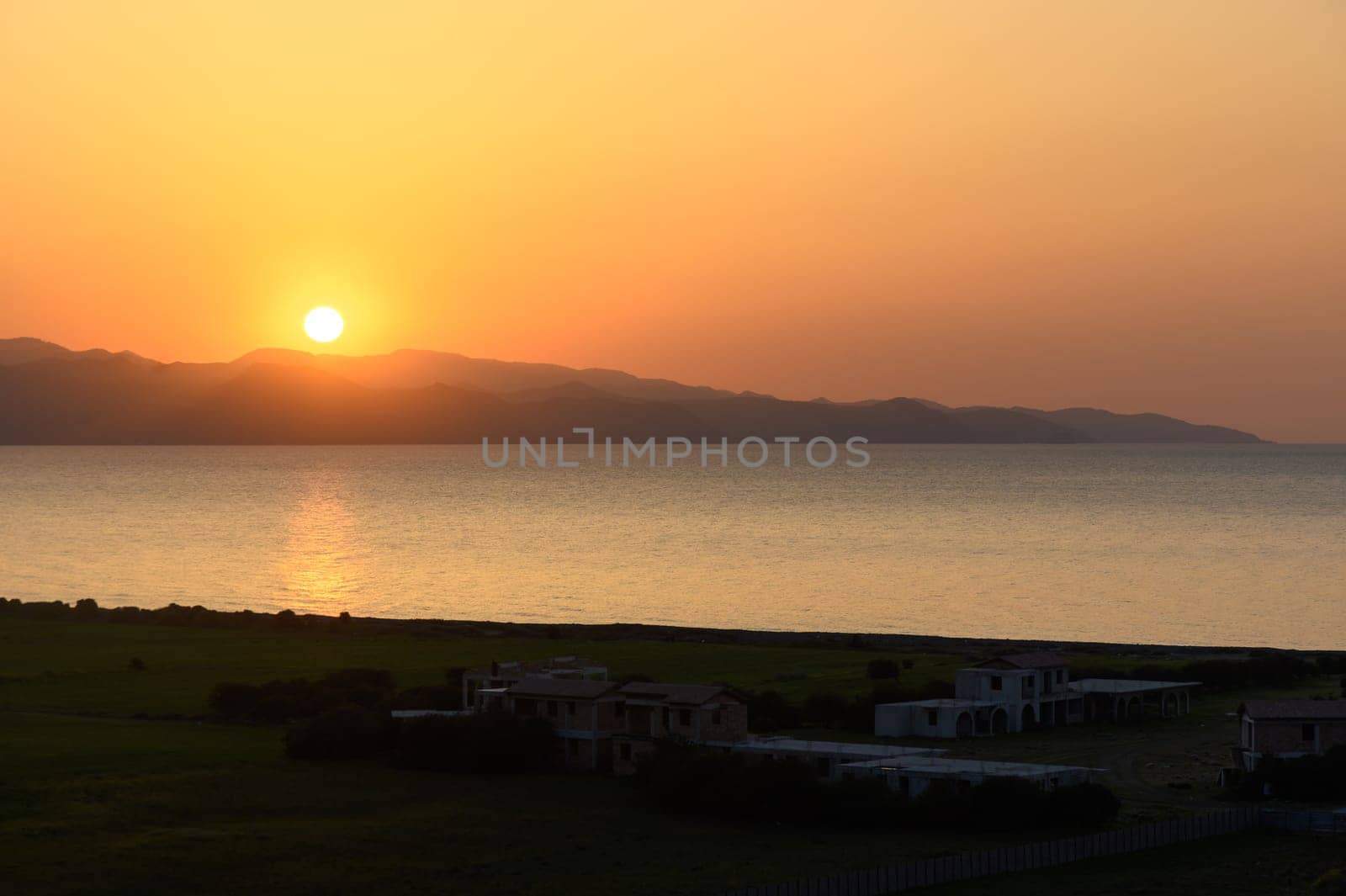 Vibrant hues of orange and yellow fill the sky as the sun dips below tranquil waters. Distant mountains create a serene backdrop, with silhouettes of houses surrounded by lush greenery, evoking peace.