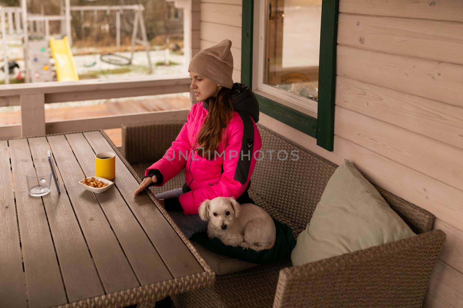 A young woman wearing warm clothing sits on a porch with her small dog beside her. She is relaxing on a cushioned bench, sipping a drink and surrounded by nature.