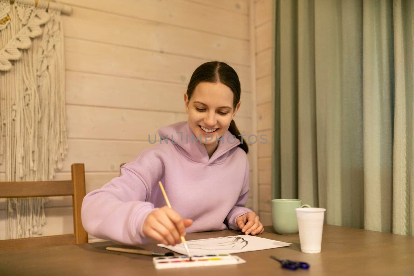 A girl with long hair smiles as she paints on a sheet of paper. She uses a brush to mix vibrant colors while seated at a wooden table. A cup of tea sits nearby, enhancing the warm atmosphere.