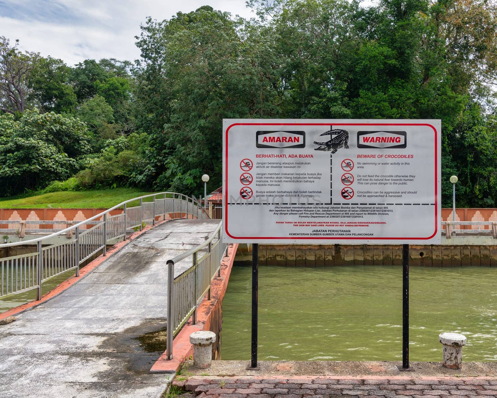 A warning sign advises about the presence of crocodiles beside a tributary of Sungai Brunei, or Brunei River in Bandar Seri Begawan. The area features a bridge and lush greenery
