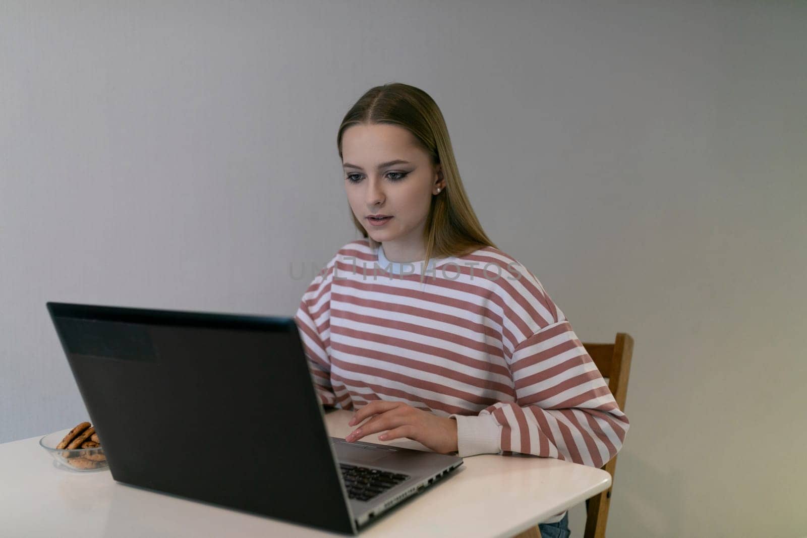 A young woman sits at a white table, deeply engaged with her laptop. Striped clothing and a plate of cookies indicate a relaxed afternoon at home.