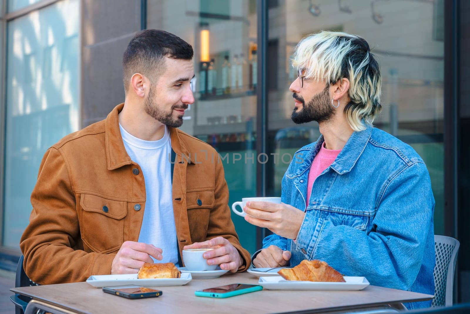 Two friends share a relaxed moment at an outdoor cafe, smiling and talking while drinking coffee and eating croissants. Casual setting with phones on table, reflecting modern urban lifestyle.