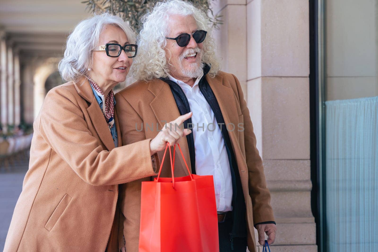 Smiling elderly couple walking outdoors, holding shopping bags while the woman points at a store window. Both wear brown coats and sunglasses, enjoying a pleasant and relaxed shopping day.