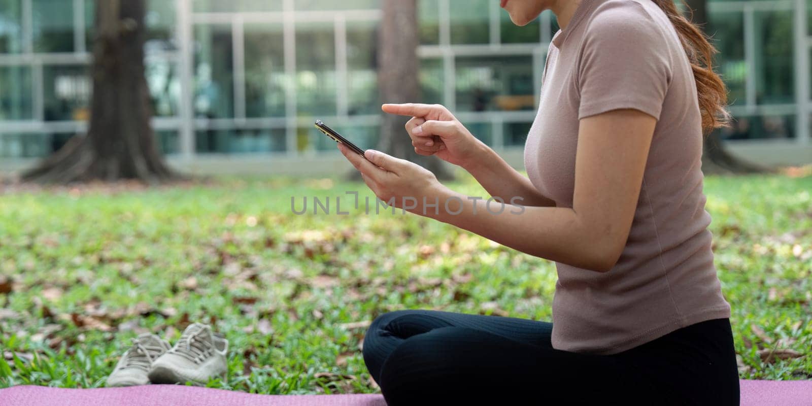 A focused woman sits cross-legged on a mat in a serene park, exploring wellness apps on her smartphone to enhance her health routine.