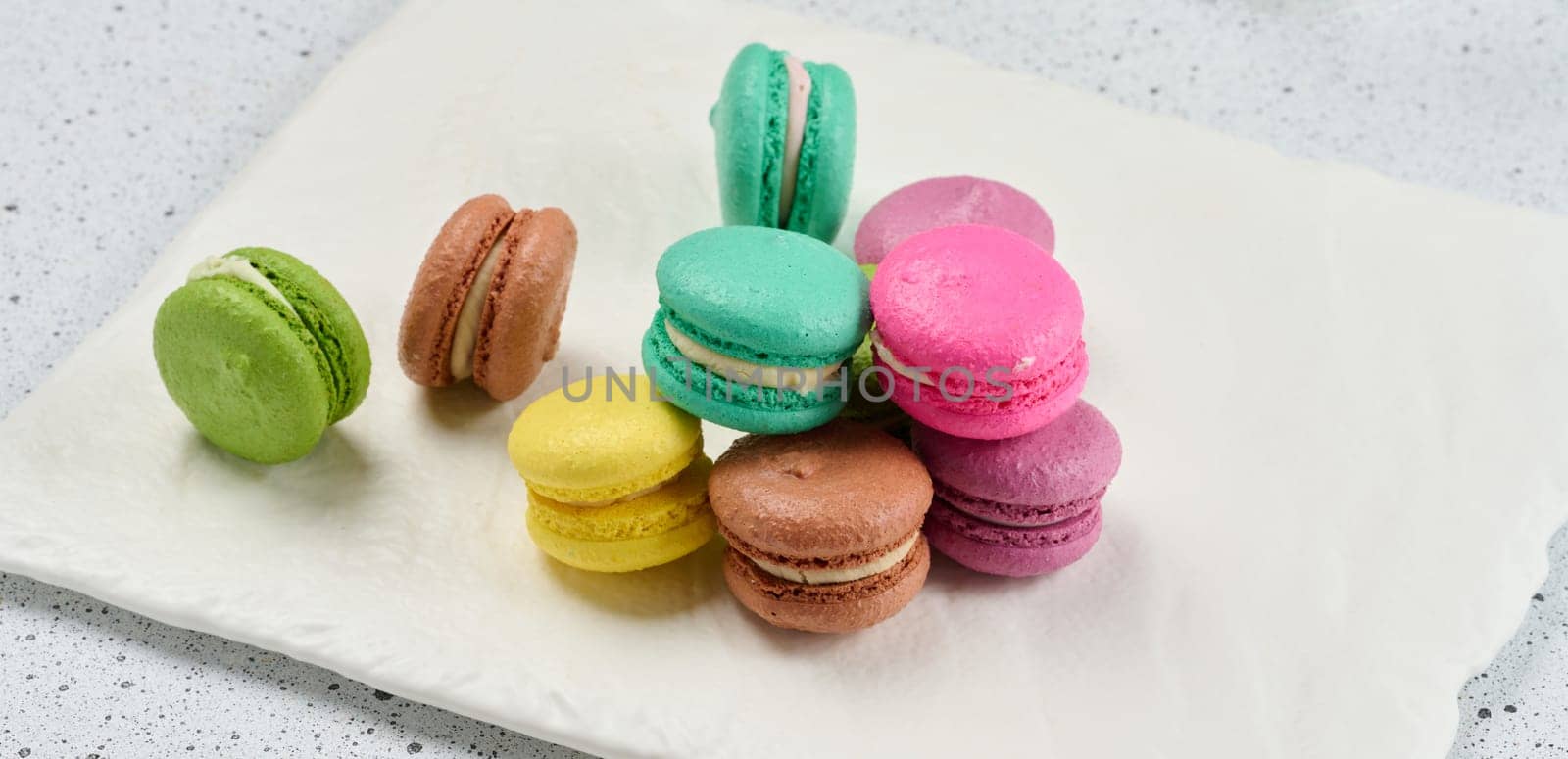 An assortment of colorful macarons on a white surface with dark speckles. The macarons have a smooth, delicate shell and a creamy filling