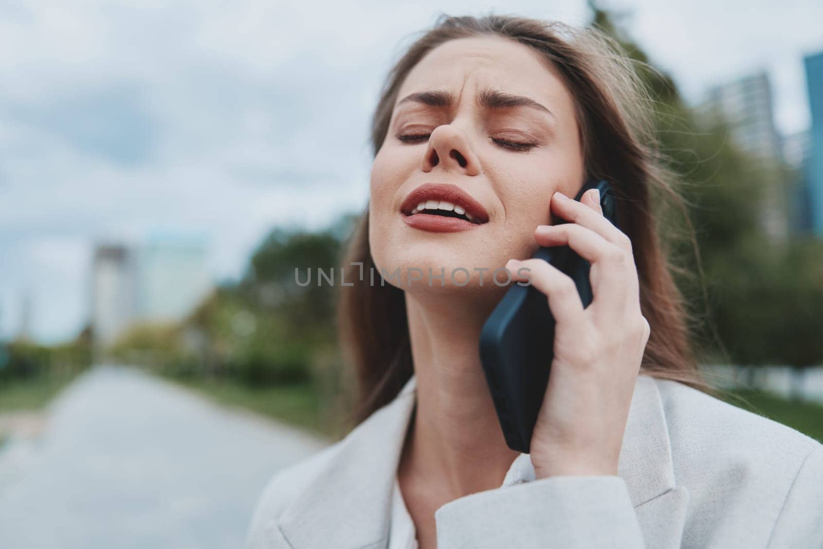 Expressive young woman in business clothing, feeling emotional while on a phone call in a city park with a clear blue sky, showcasing the challenges of modern professional life.