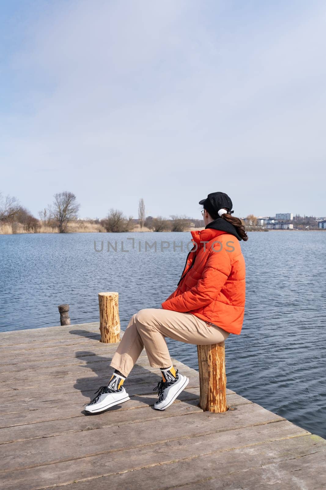 A young woman in an orange jacket sits on a wooden pier, gazing out at a tranquil lake on a sunny day.