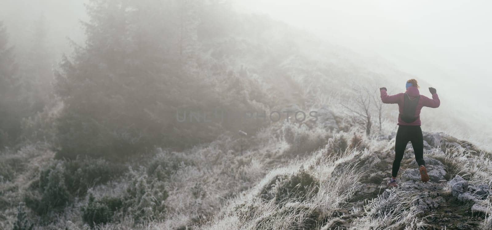 A woman athlete running along a rocky mountain trail in cold, misty weather, wearing a warm jacket, gloves, and a headband, showcasing endurance and determination.