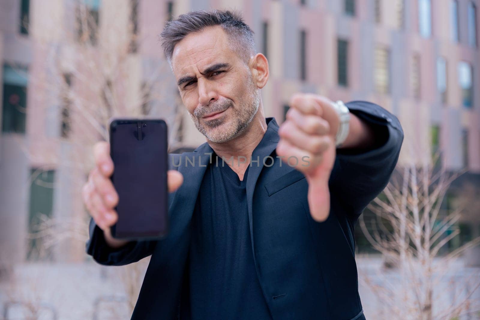 Businessman in black outfit shows his smartphone and gives a thumbs down He serious confidently in an urban background, suggesting success and desapproval of modern technology looking at camera