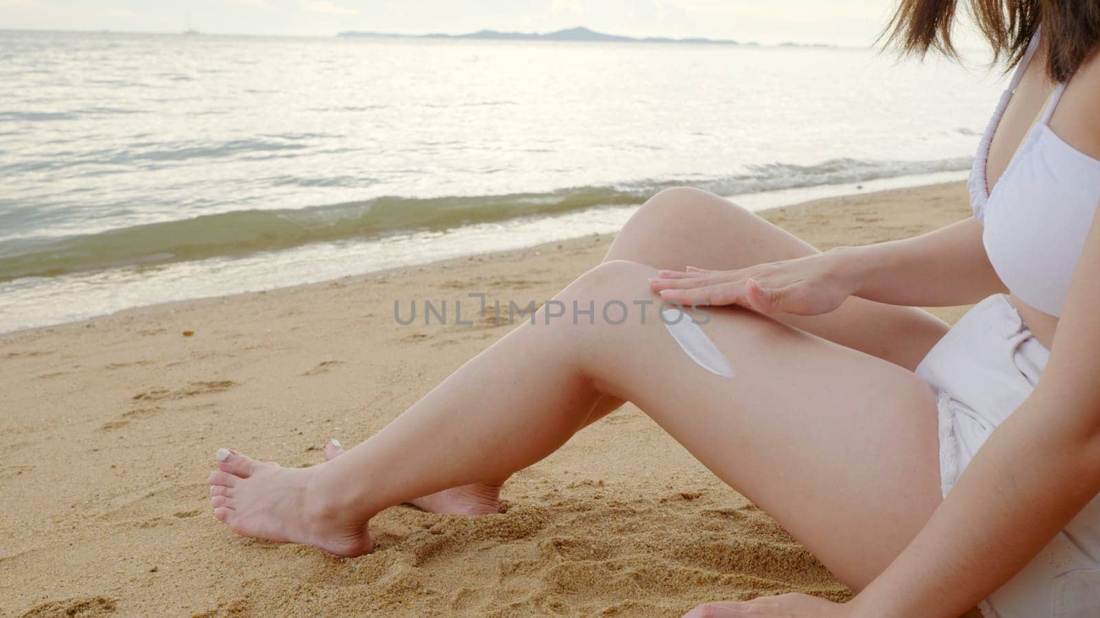 Beautiful Asian woman apply sun protection cream on tanned on leg at tropical beach with white sand, Sun Cream Skincare, Close up female in bikini using sunscreen lotion on back body before sunbathing
