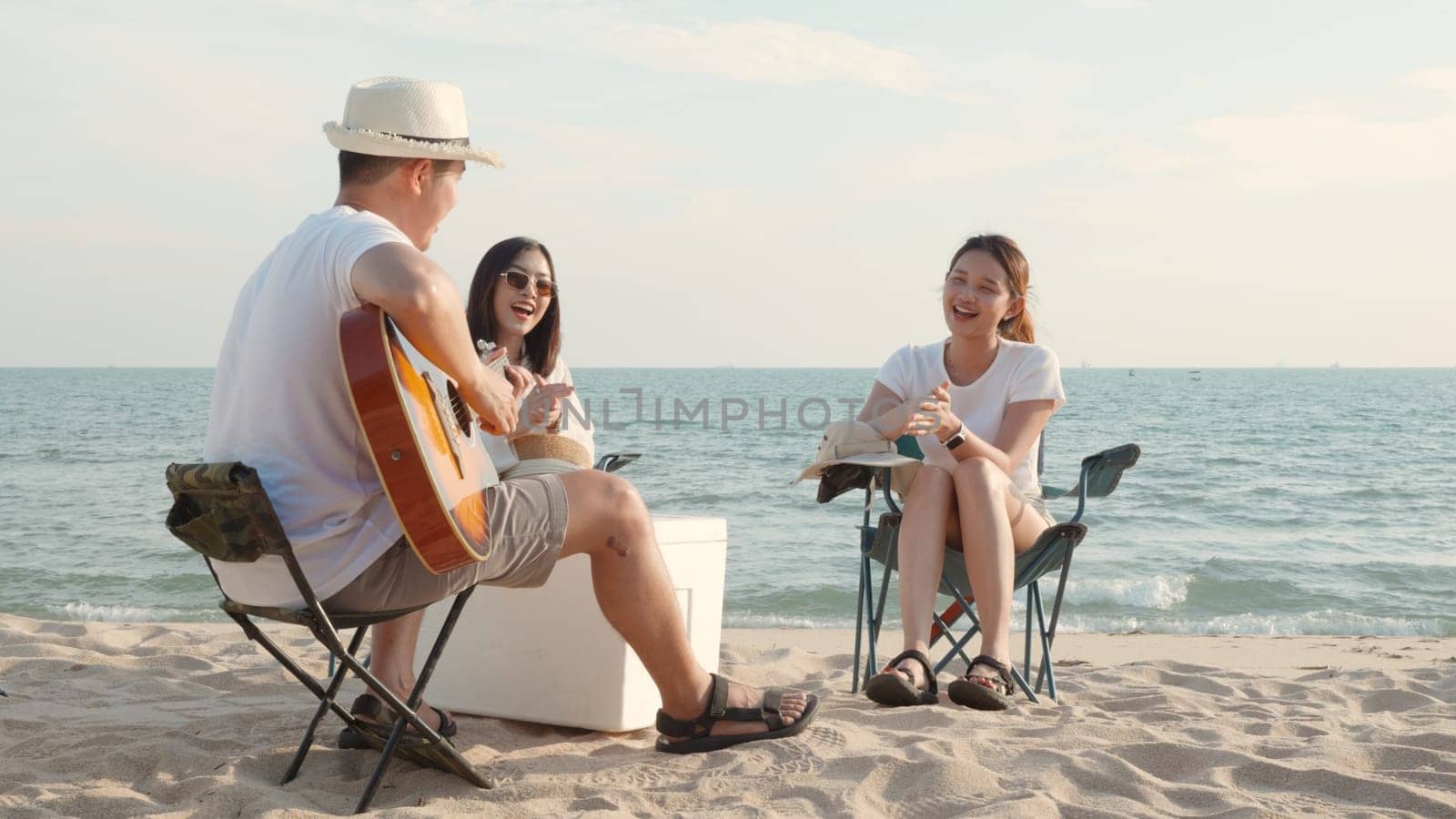Happy friend have fun playing guitar and clap in camp they smiling together in holiday on sand beach near camping tent vacation time at sunset, Young Asian group woman and man in summer travel outdoor