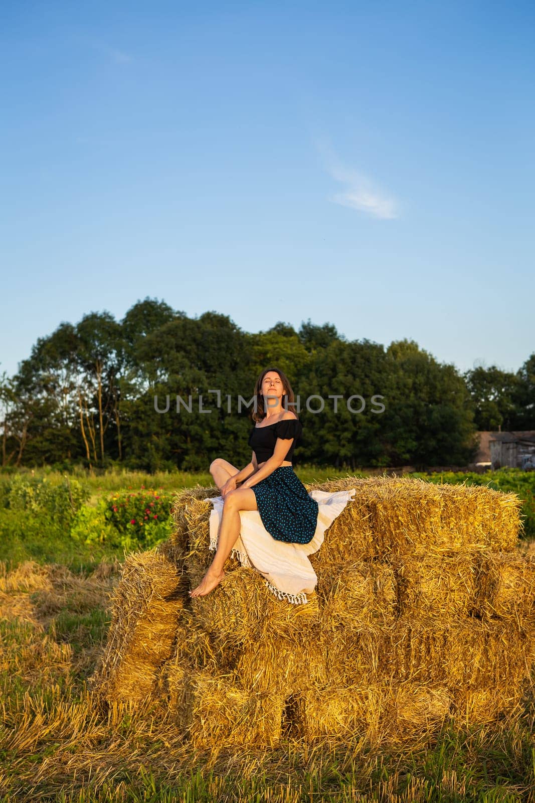 Young beautiful woman sits on a large pile of straw bales. Freedom, wind, setting sun