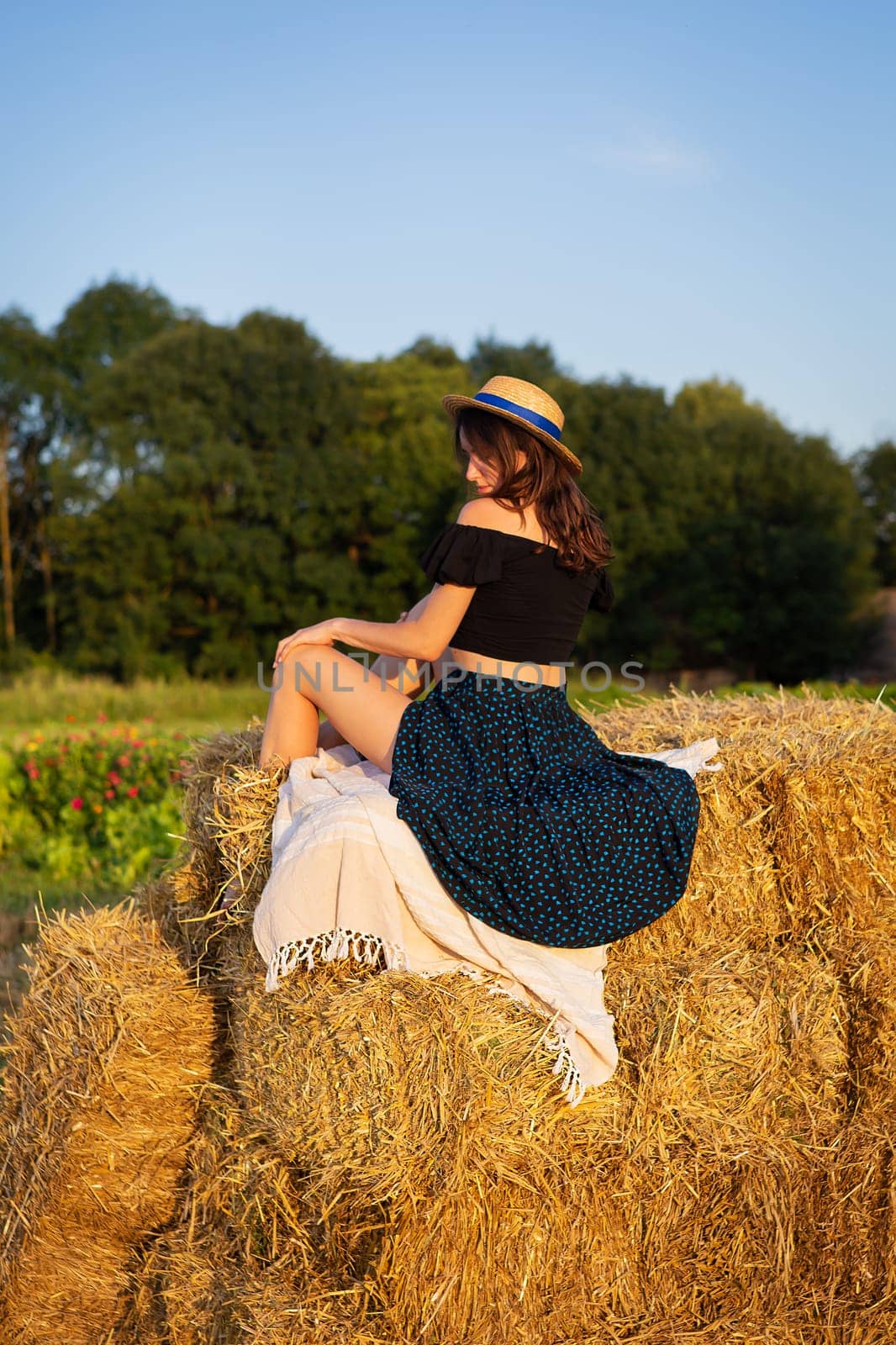 Young beautiful woman in a straw hat looks at the setting sun, the girl sits on a large pile of straw bales. Warm summer evening