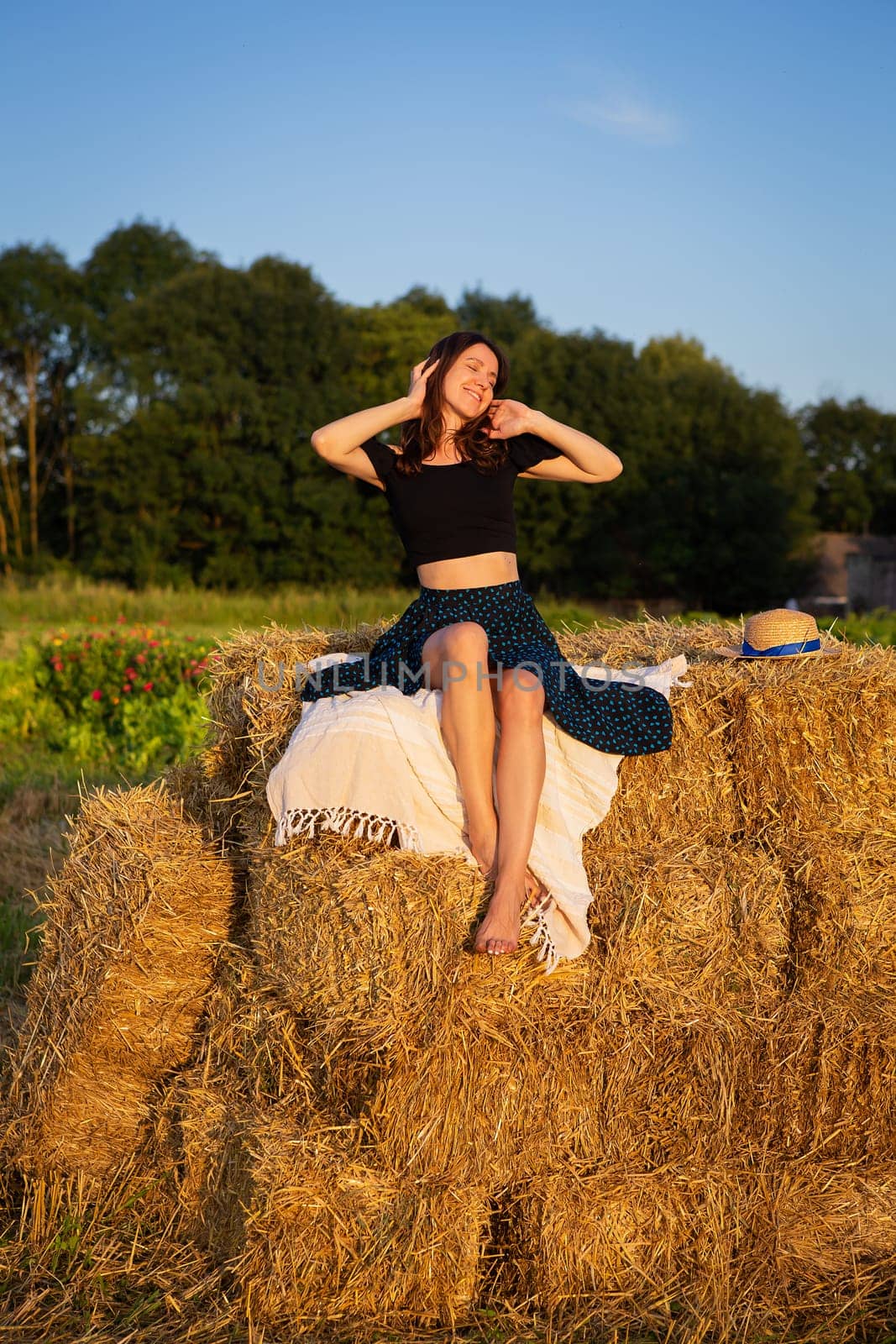 Young beautiful woman looks at the setting sun, sits on a large pile of straw bales