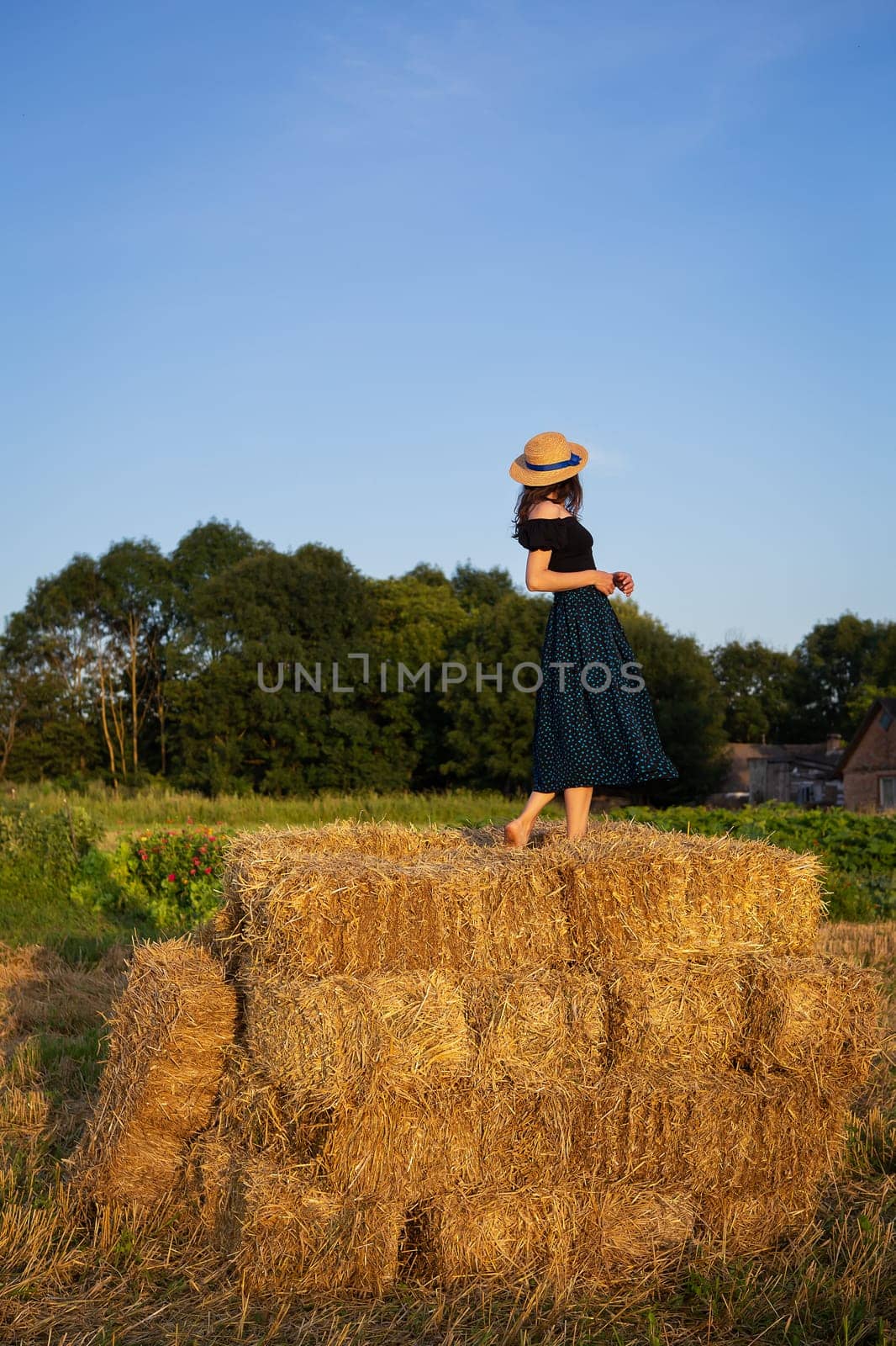 A young beautiful woman in a straw hat stands on a large pile of straw bales. Freedom, wind, setting sun