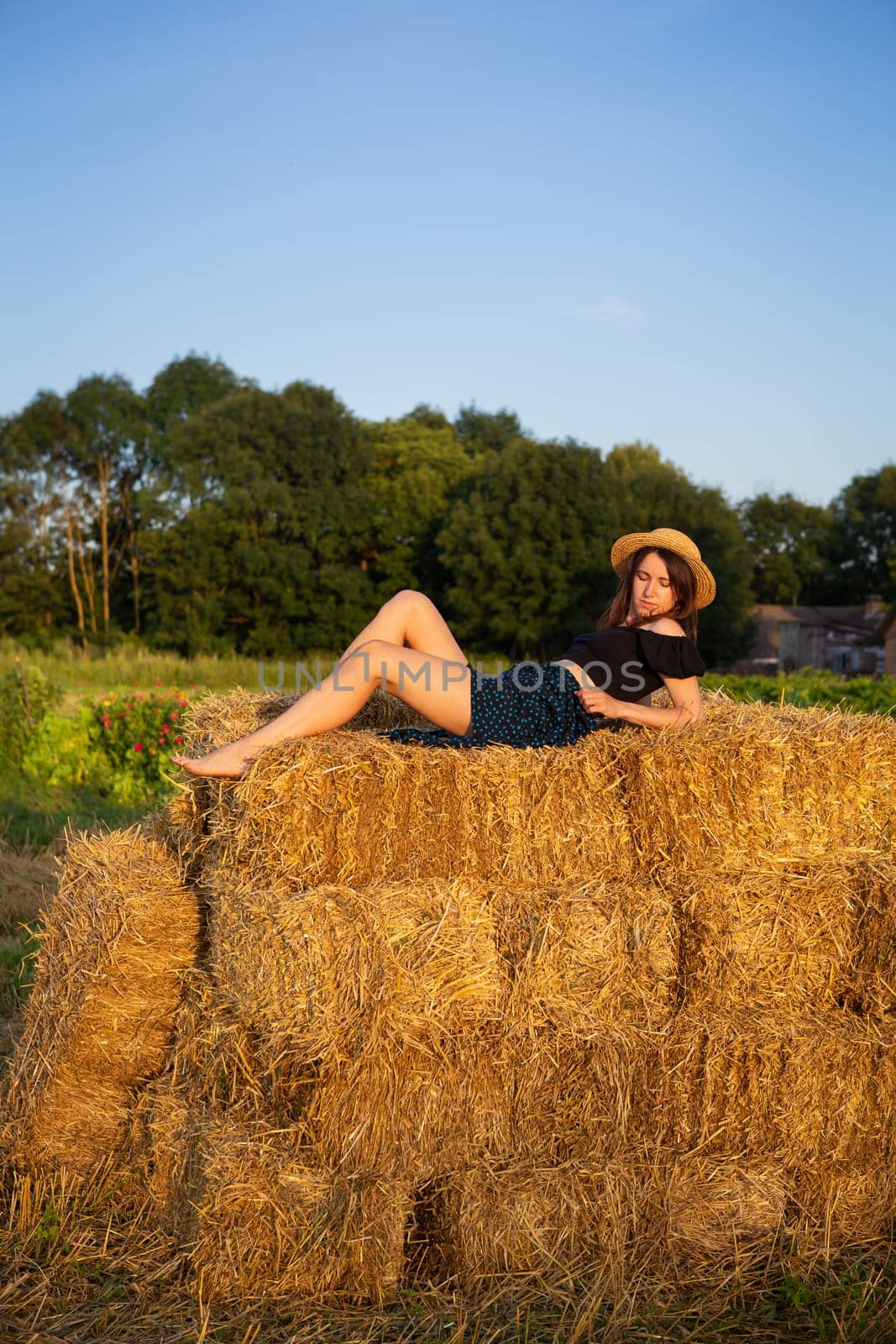 A young beautiful woman in a straw hat lies on a large pile of straw bales and looks at the setting sun