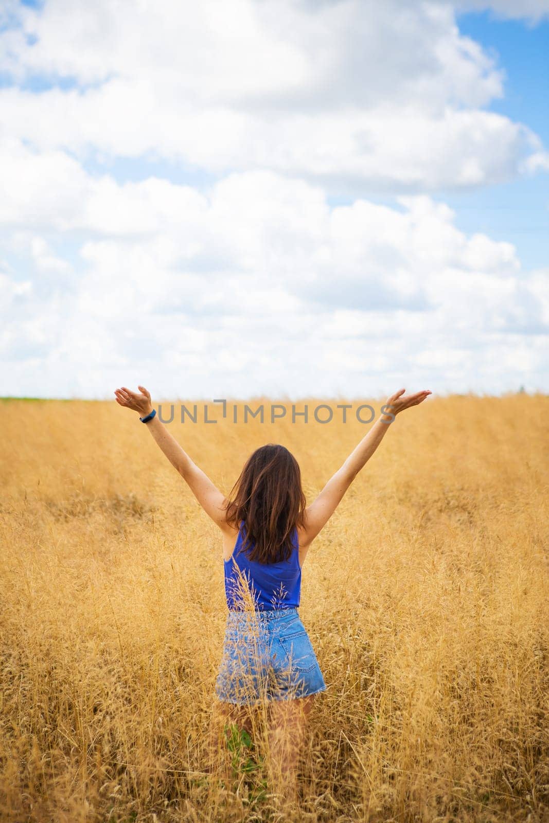 Portrait of a young beautiful brunette woman in a blue T-shirt and denim shorts stands in the middle of the field, exposing her face to the sun on the background of a summer day outdoors