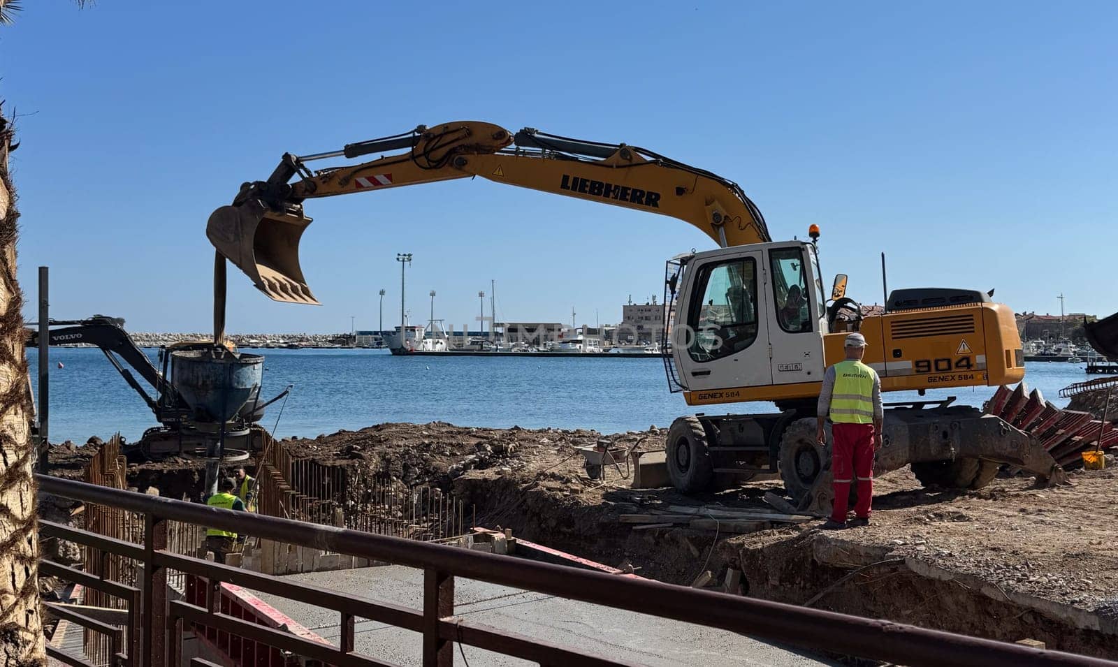 Budva,Montenegro-April 11,2025:Liebherr and Volvo excavators at a waterside construction site with workers and a marina in the background. High quality photo