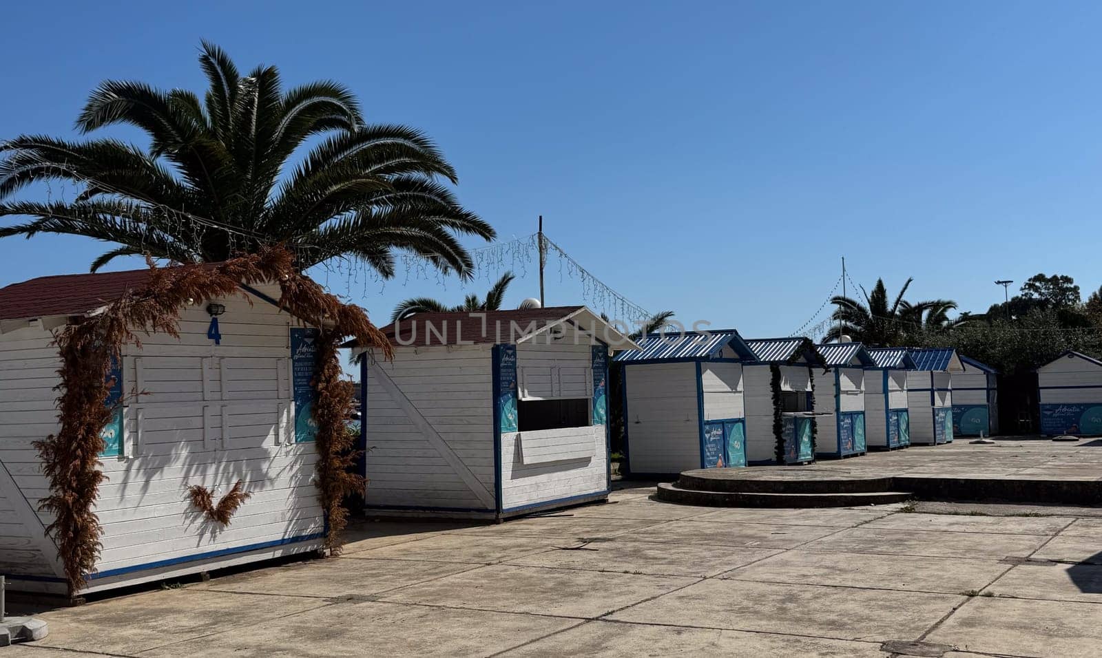 Budva,Montenegro-April 11,2025:Row of small, closed white wooden beach market huts with palm trees and string lights on a sunny day.