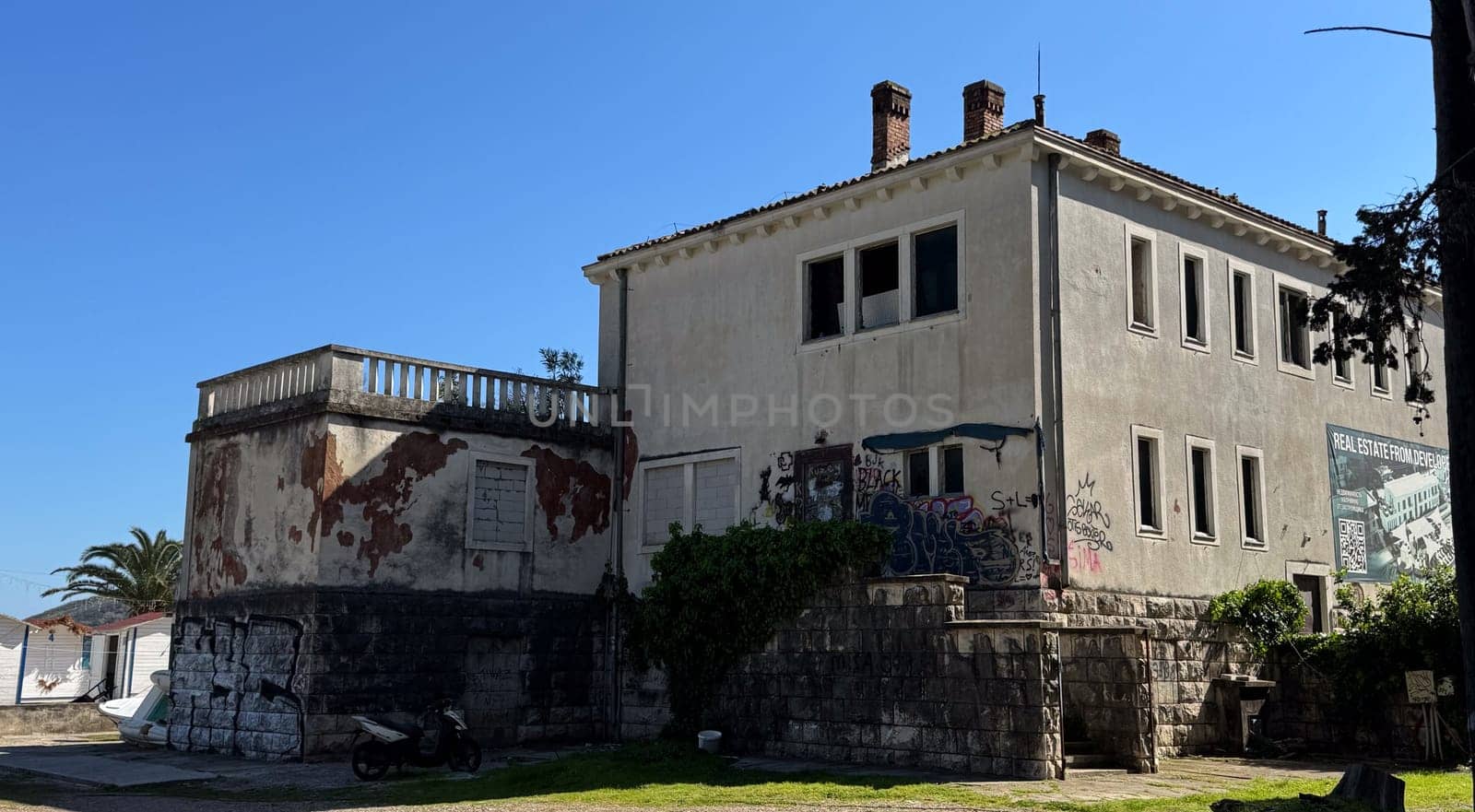 Budva,Montenegro-April 11,2025:Old two-story building with peeling paint, graffiti, and boarded windows under clear sky, surrounded by palm trees.