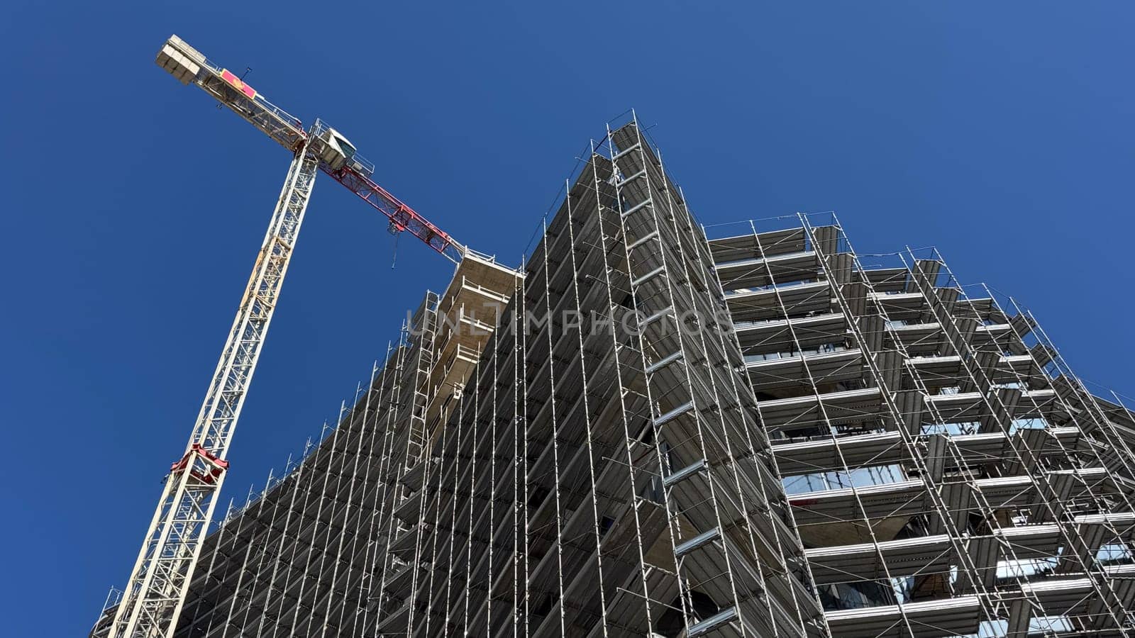 Budva, Montenegro-April 11,2025:A modern high-rise with curved facade surrounded by scaffolding and a nearby tower crane, under clear sky.