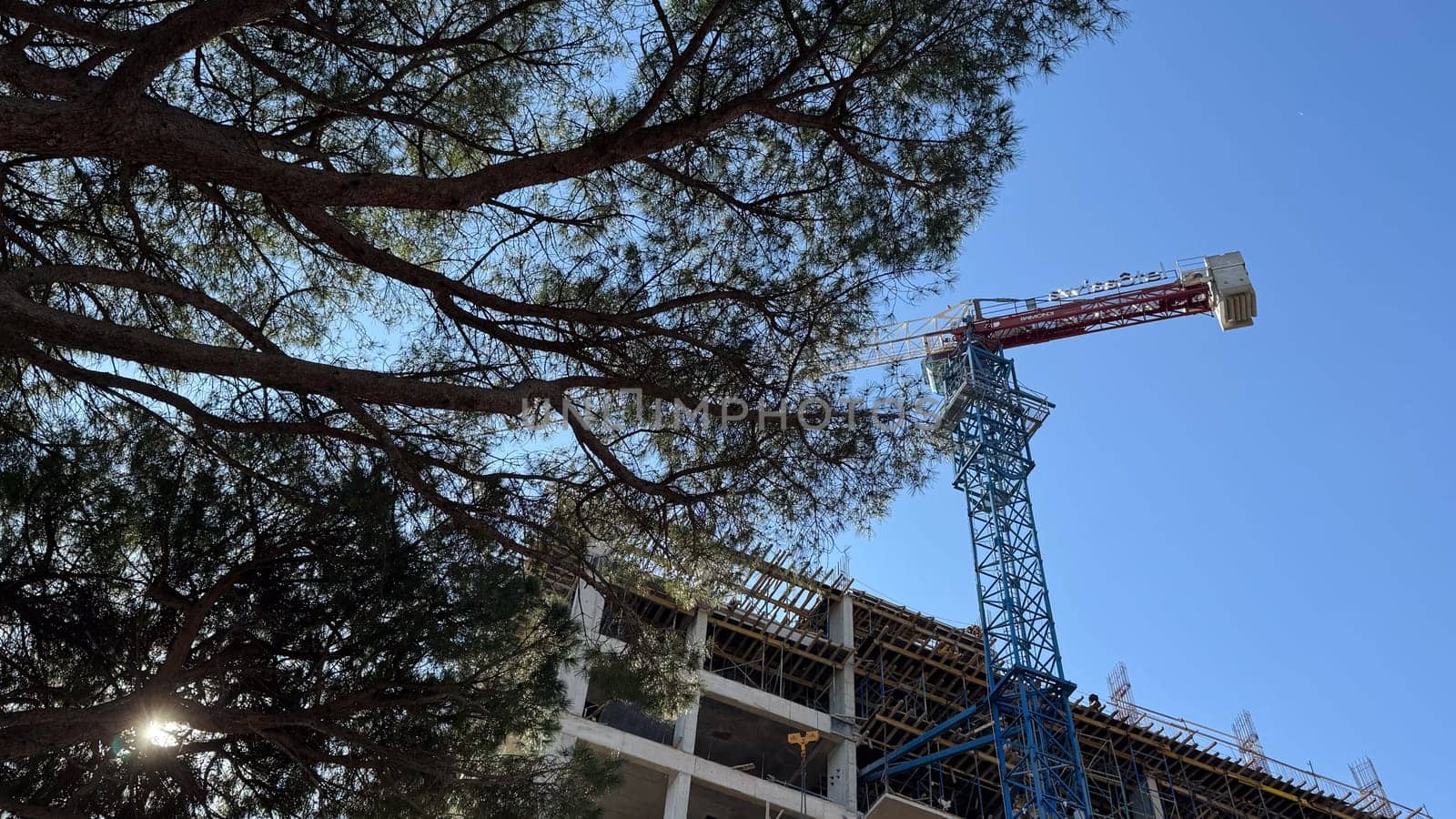 Budva,Montenegro-April 11,2025:A tall construction Blue tower crane with the SWISSOTEL sign towers over a multi-story building under construction framed by tree branches. High quality photo