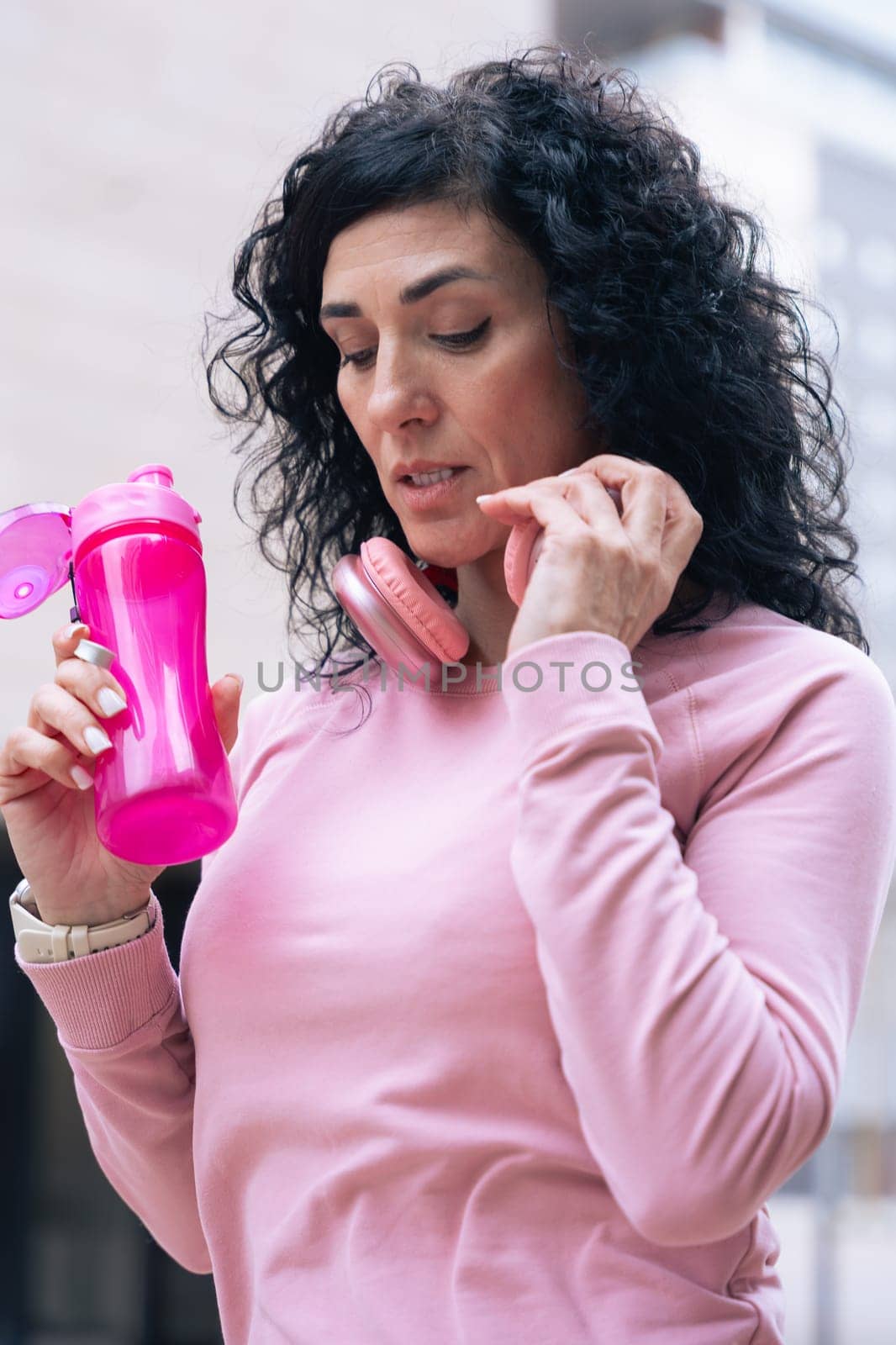 Caucasian athletic woman wearing a pink hoodie and headphones around her neck holds a pink water bottle. She stands in an urban setting, appearing thoughtful and focused after a workout. Vertical