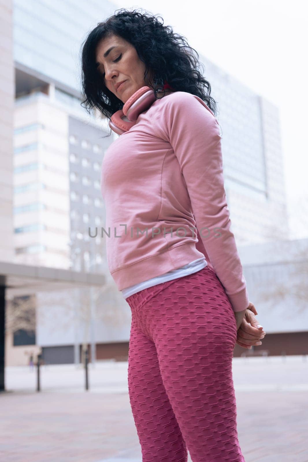 Caucasian adult woman with curly black hair stretching her arms outdoors in an urban setting.she is wearing a pink sweatshirt and exercising for a healthy lifestyle, fitness, and wellness in the city