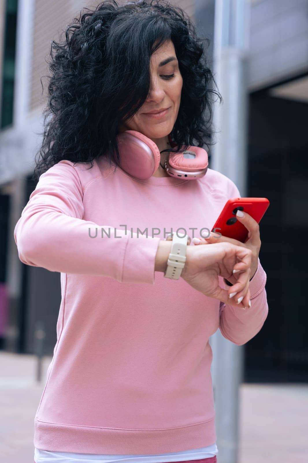 Middle adult Caucasian woman with curly black hair checking her smartwatch outdoors. She is wearing a pink sweatshirt and pink headphones, representing fitness, technology, and an active lifestyle.