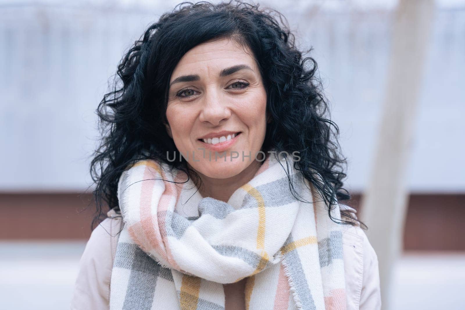 Horizontal portrait of a woman sitting on a bench outdoors. She wears a beige coat and scarf, looking calm and confident. Urban background with winter atmosphere and seasonal fashion concept.