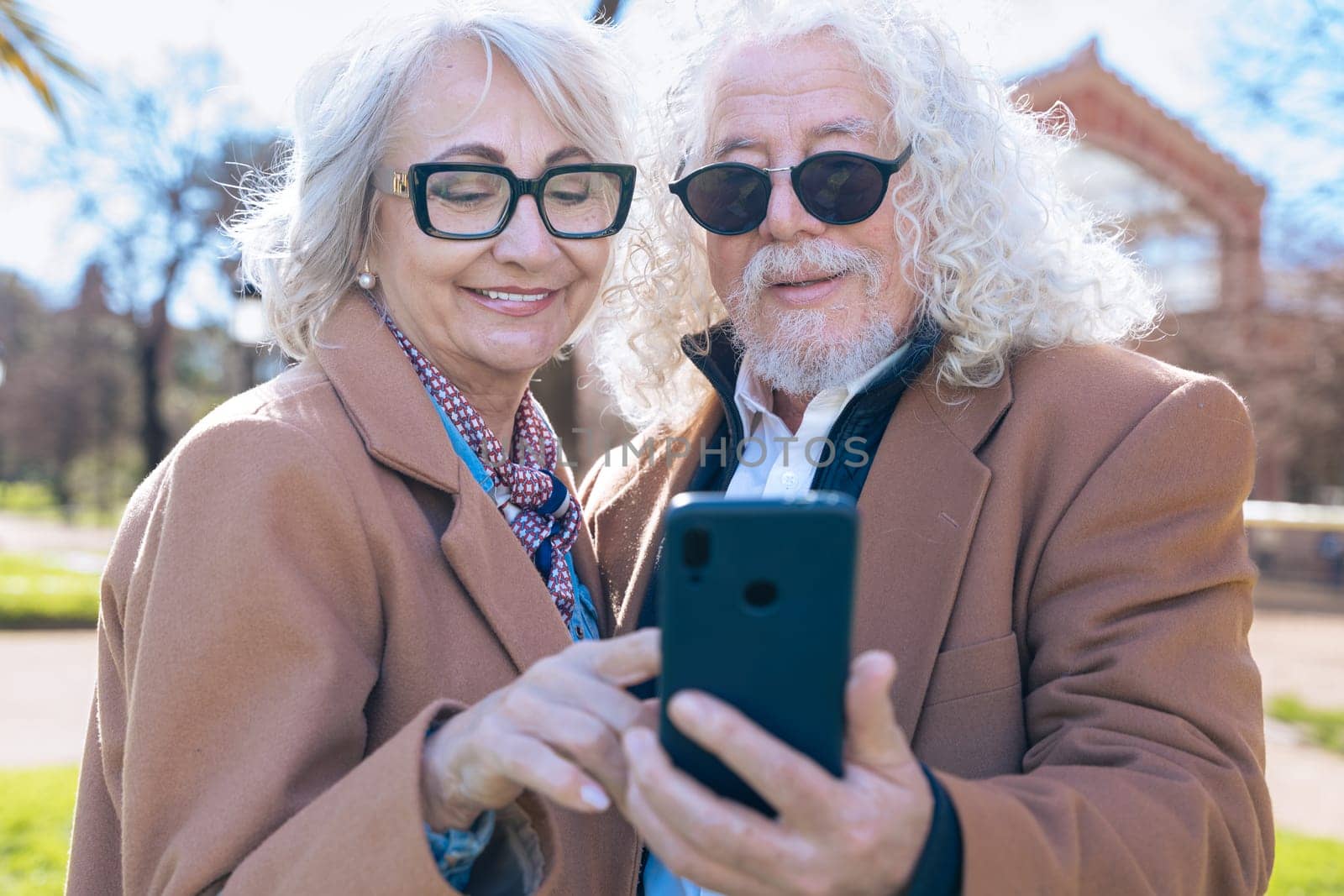 Caucasian elderly couple standing outdoors using a smartphone app to check social media. They smile while sharing the screen, enjoying a sunny day together in casual brown jackets and glasses.