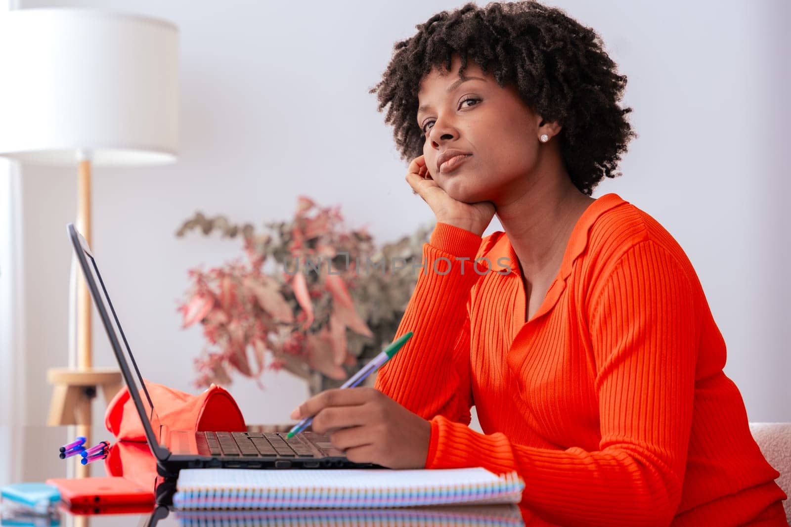 African American woman sitting at home, thinking while working on her laptop. She holds a pen and looks away thoughtfully. Concept of remote work, education, focus, and digital lifestyle.