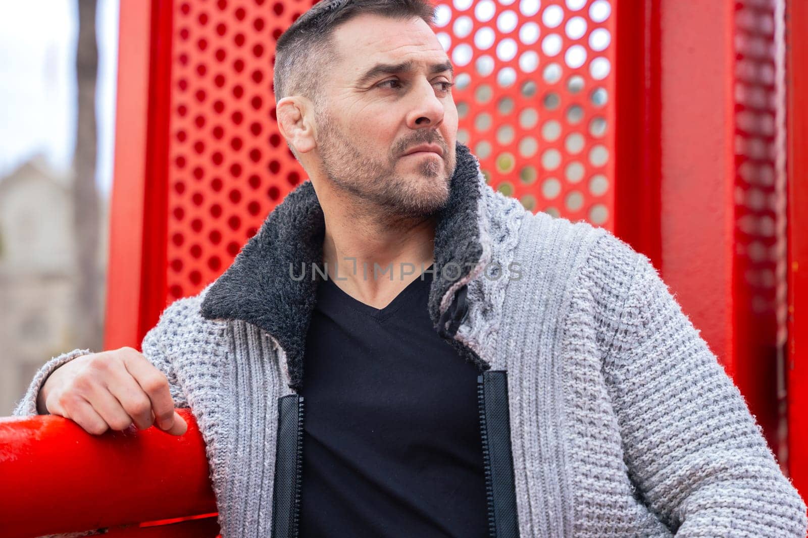 Portrait of a serious caucasian man standing by a bold red metal structure in an urban environment. Wearing a gray sweater and black shirt, he looks confidently at the camera with city view behind