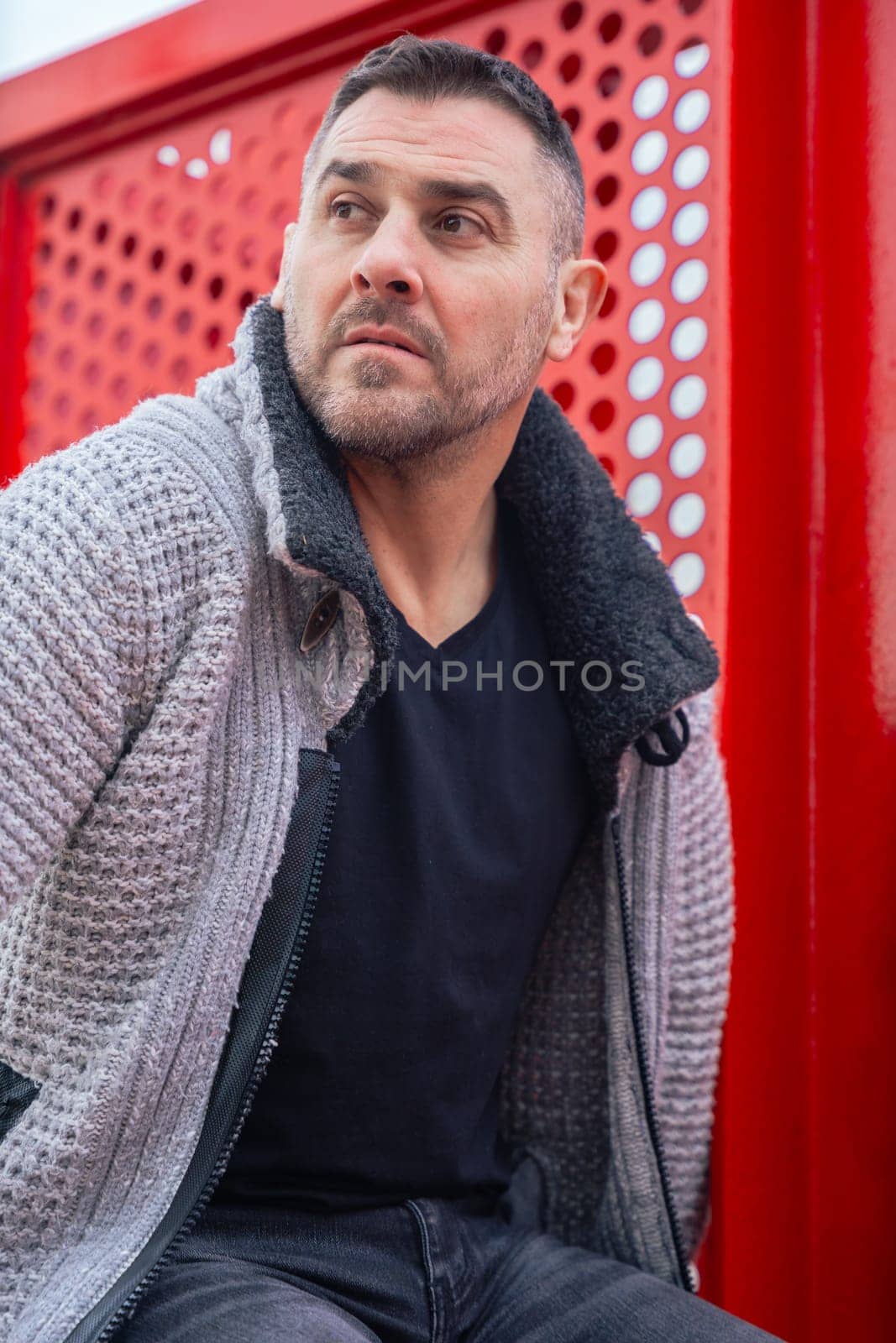 Portrait of a serious caucasian man standing by a bold red metal structure in an urban environment. Wearing a gray sweater and black shirt, he looks confidently at the camera with city view behind