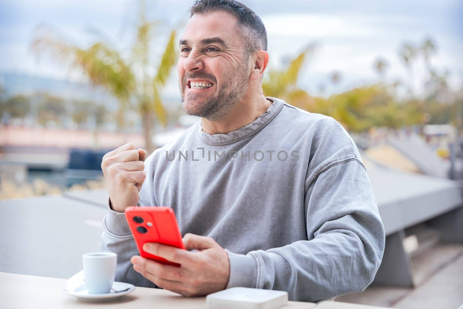 Excited caucasian man reacts with joy after winning an online game bet on his smartphone, sitting at an outdoor cafe table with coffee, enjoying his moment of triumph and happiness