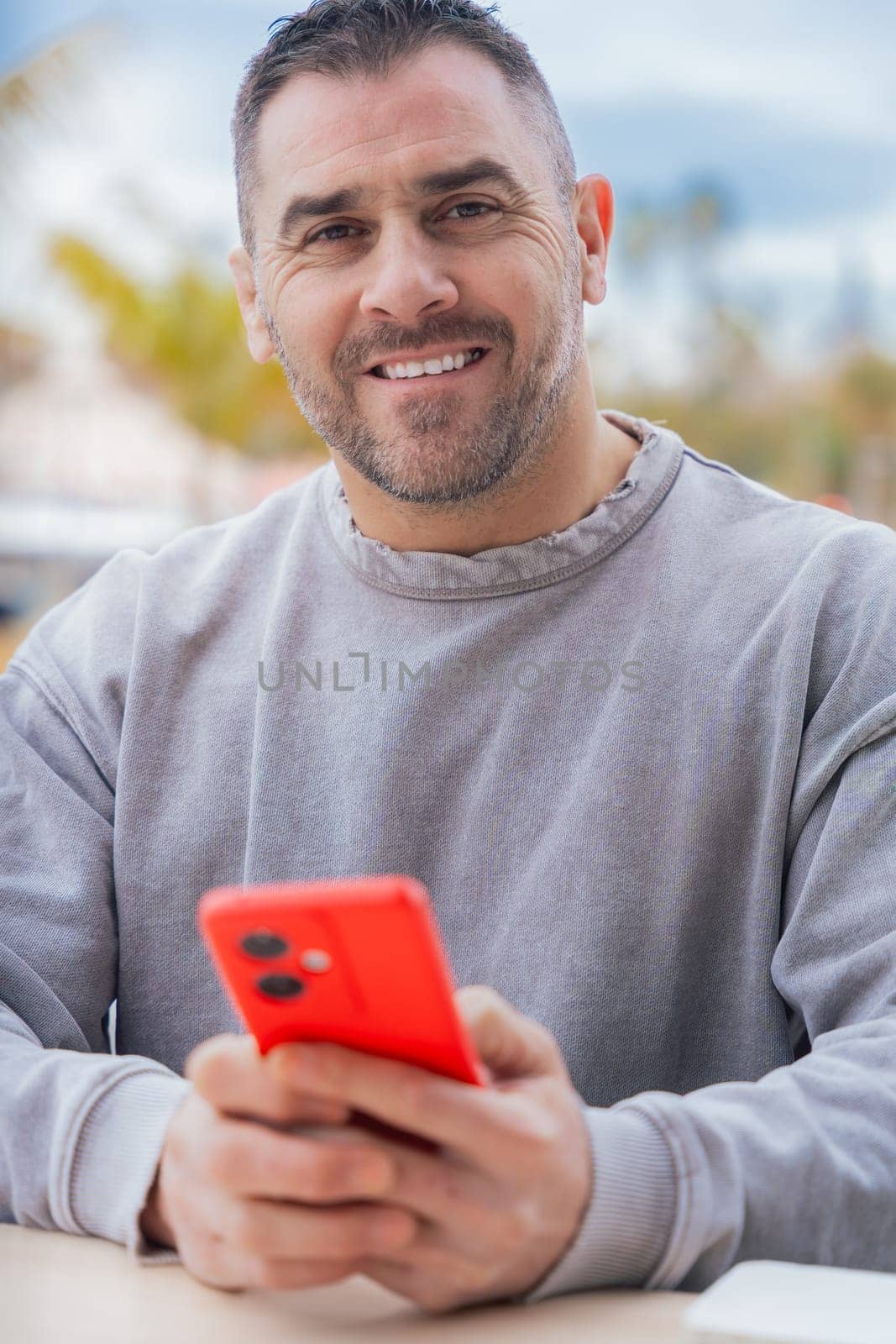 Caucasian man sitting at an outdoor cafe table, smiling at the camera while holding a red smartphone. Casual outfit and relaxed expression create a warm, approachable and modern tech vibe.vertical